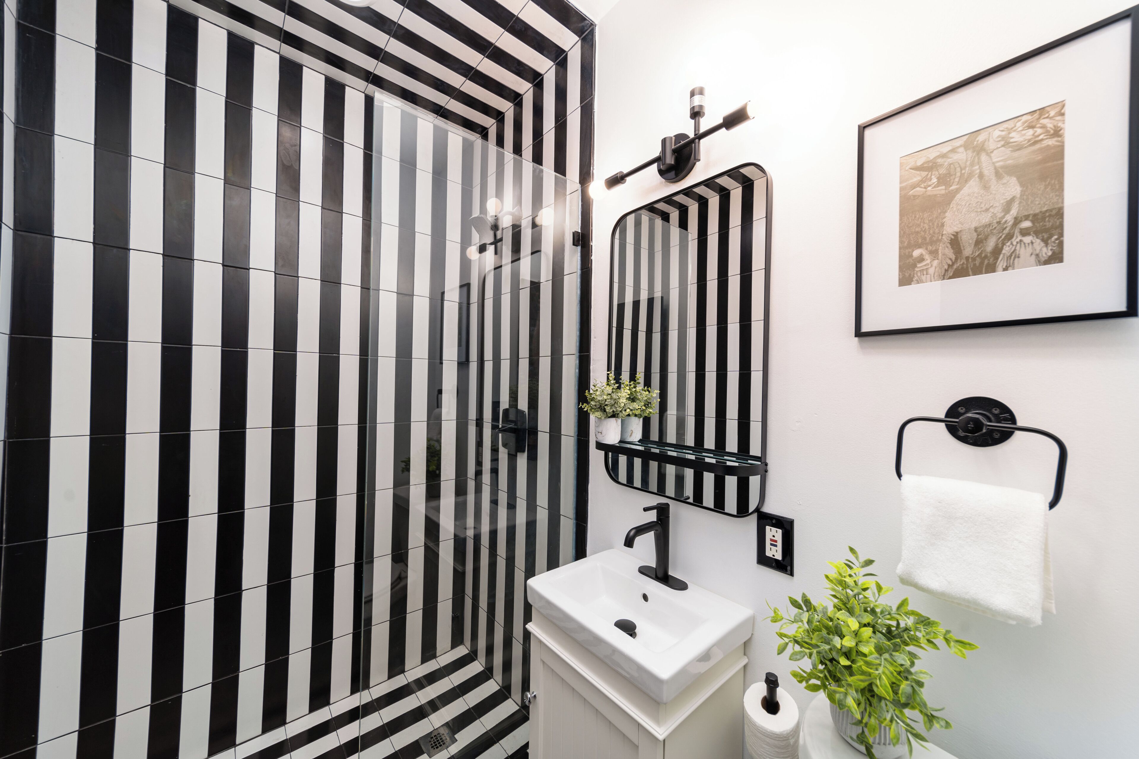 Une salle de bain avec un mur recouvert de carreaux noirs et blancs formant des rayures, un lavabo blanc, un miroir, un porte-serviettes et un tableau encadré accroché au mur.