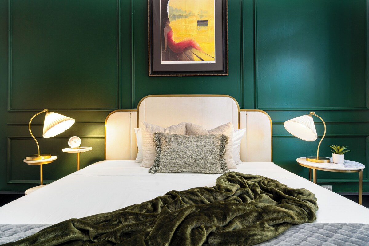 A bedroom with a bed, two bedside lamps, a framed painting, and a green wall.