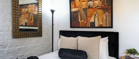 Un dormitorio moderno con cama, un cuadro abstracto grande, un espejo con marco decorativo y una mesita de noche con un libro y una planta.
