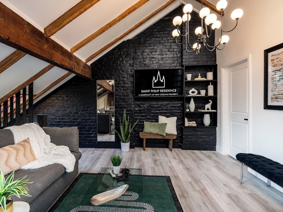 A loft-style living room with exposed wooden beams, a black brick wall, a chandelier, and a view into the kitchen.