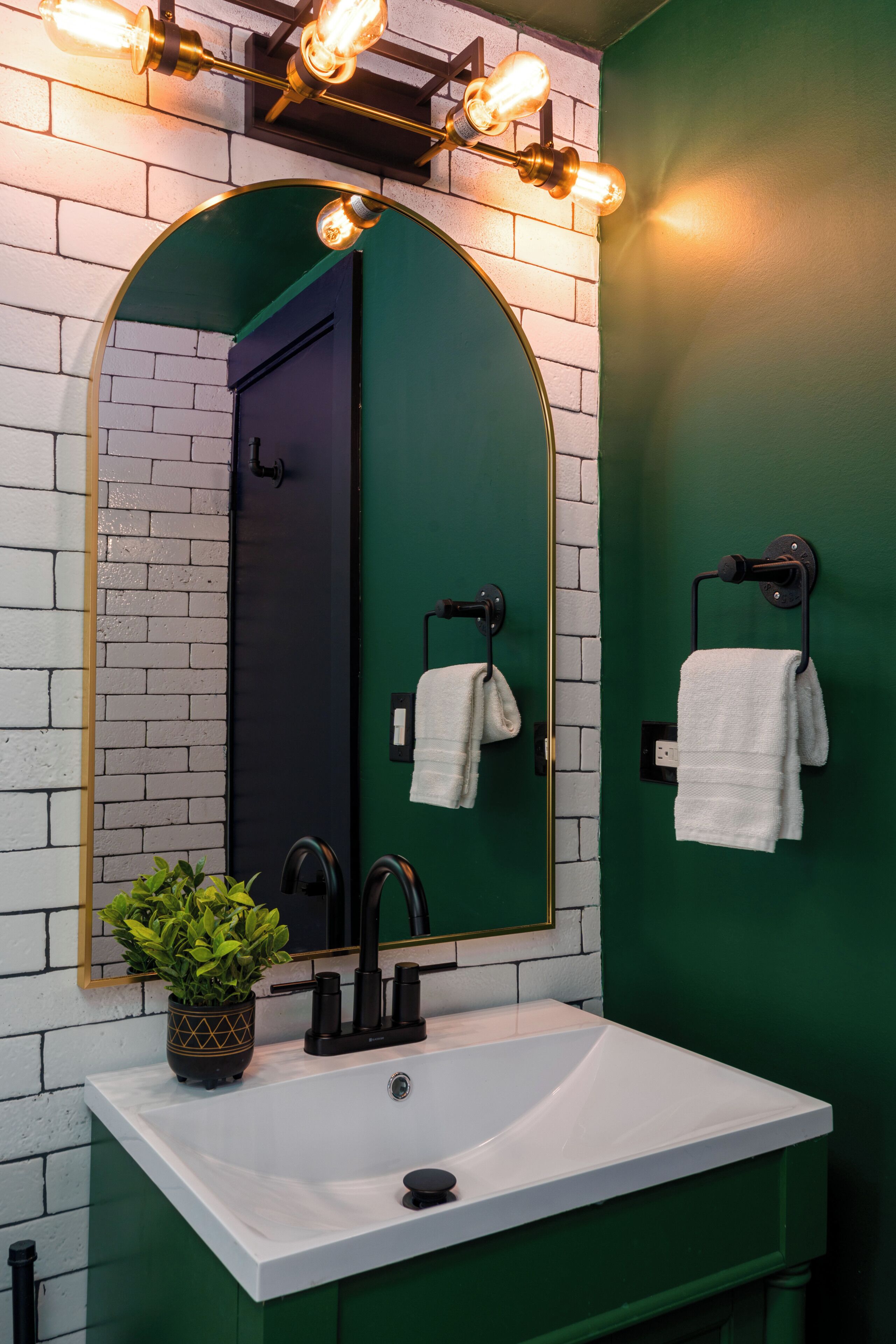 Un baño con un lavabo blanco, un espejo con luces, una pared verde y dos toallas colgadas de ganchos.