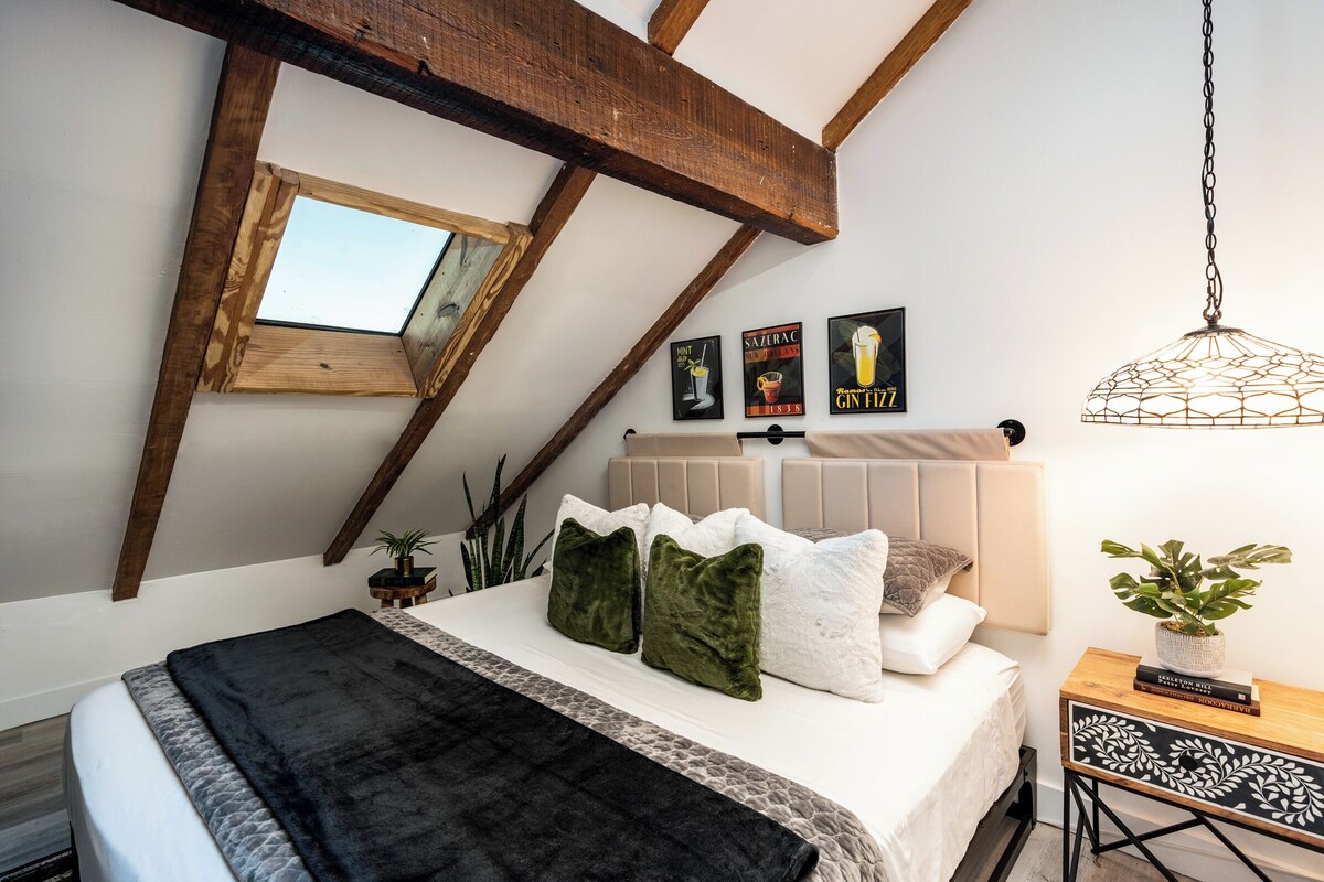 A bedroom with a sloped ceiling, a skylight, a bed with multiple pillows, a bedside table with books, and wall art.
