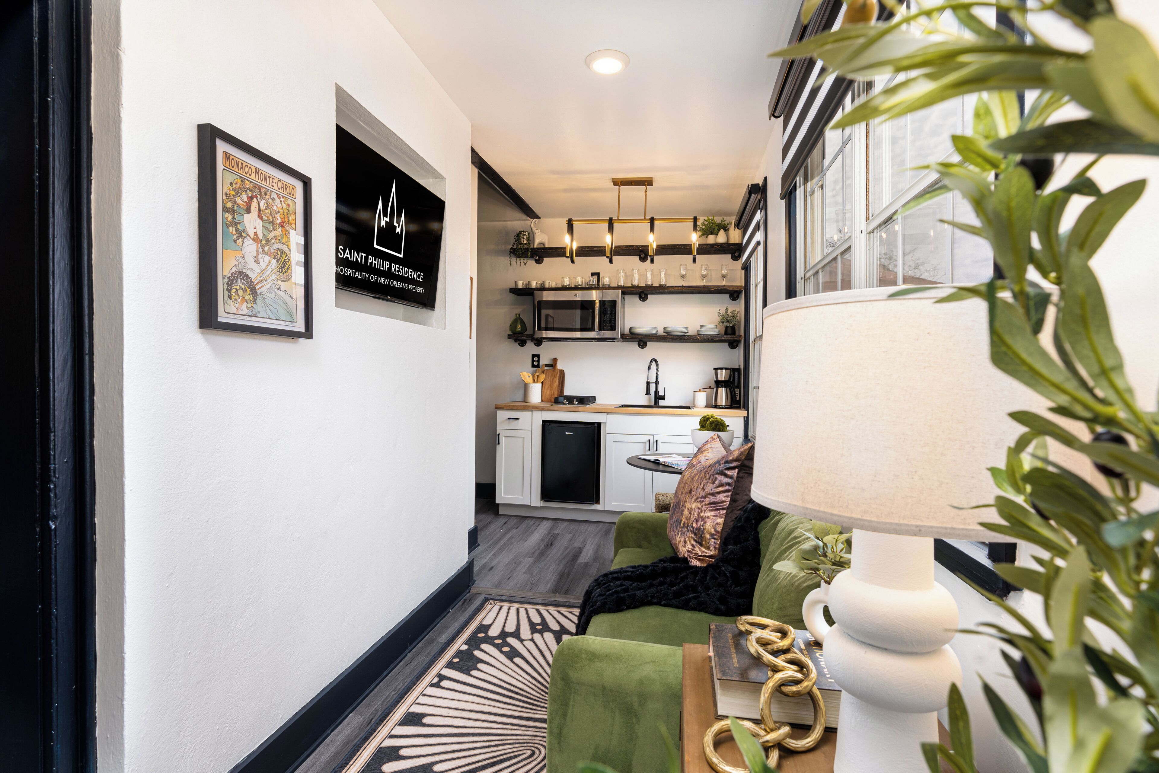 A modern kitchen with white cabinets, a black refrigerator, and a microwave. There is a dining area with a green sofa and a lamp.