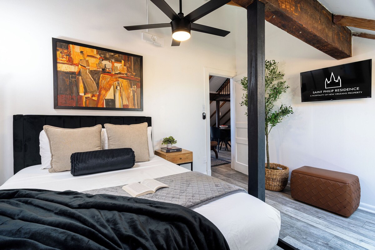 A bedroom with a bed, a ceiling fan, a painting, a television, and a plant.