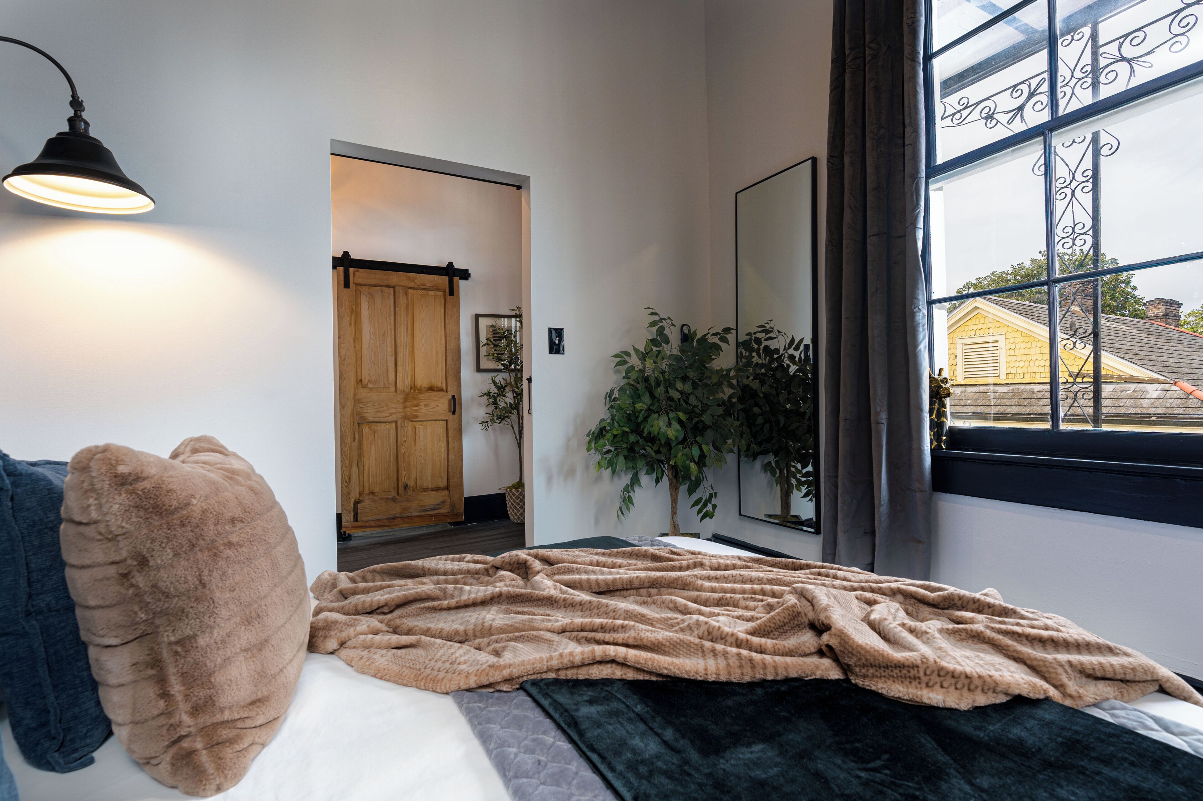Un dormitorio con una cama, una ventana con cortinas, una planta y una puerta que da a otra habitación.