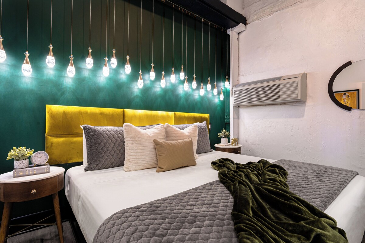 A bedroom with a bed, pillows, a bedside table with a clock, a potted plant, and a wall-mounted air conditioner.