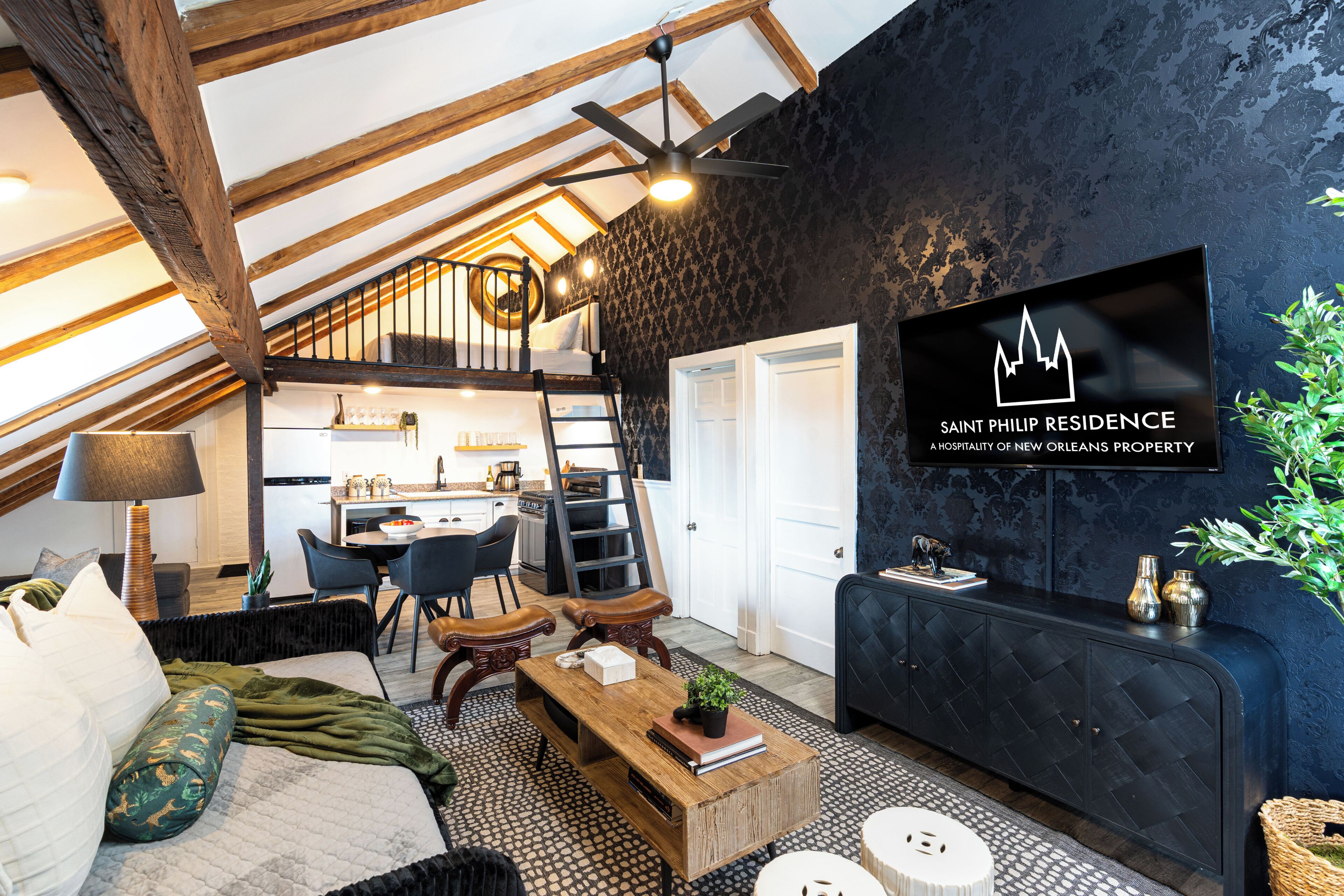 Un salón estilo loft con techo alto, vigas de madera a la vista, una pared de acento negra, un televisor y una cocina al fondo.