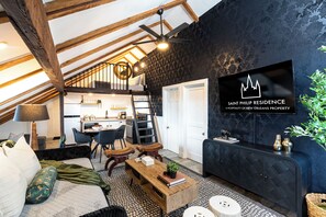 A loft-style living room with a high ceiling, exposed wooden beams, a black accent wall, a television, and a kitchen in the background. - Saint Philip French Quarter (New Orleans)