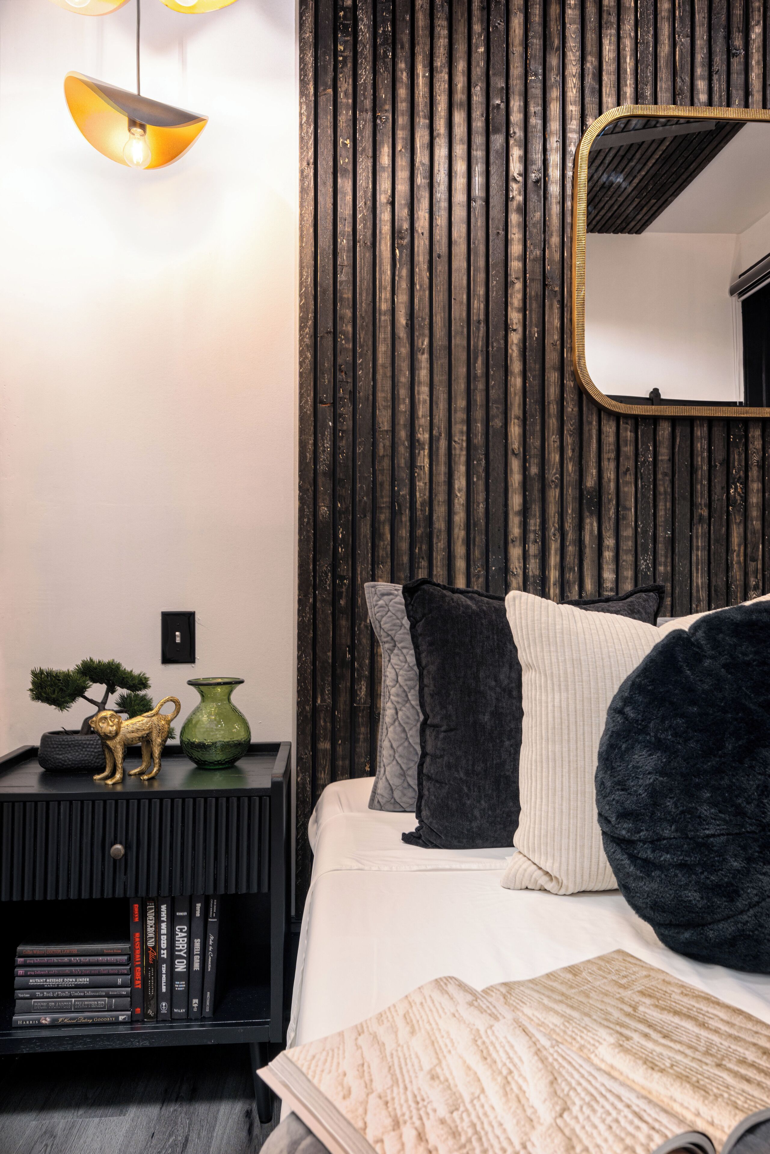 Quarto com cama, almofadas, mesa de cabeceira com livros, um vaso verde e uma pequena estatueta de macaco.