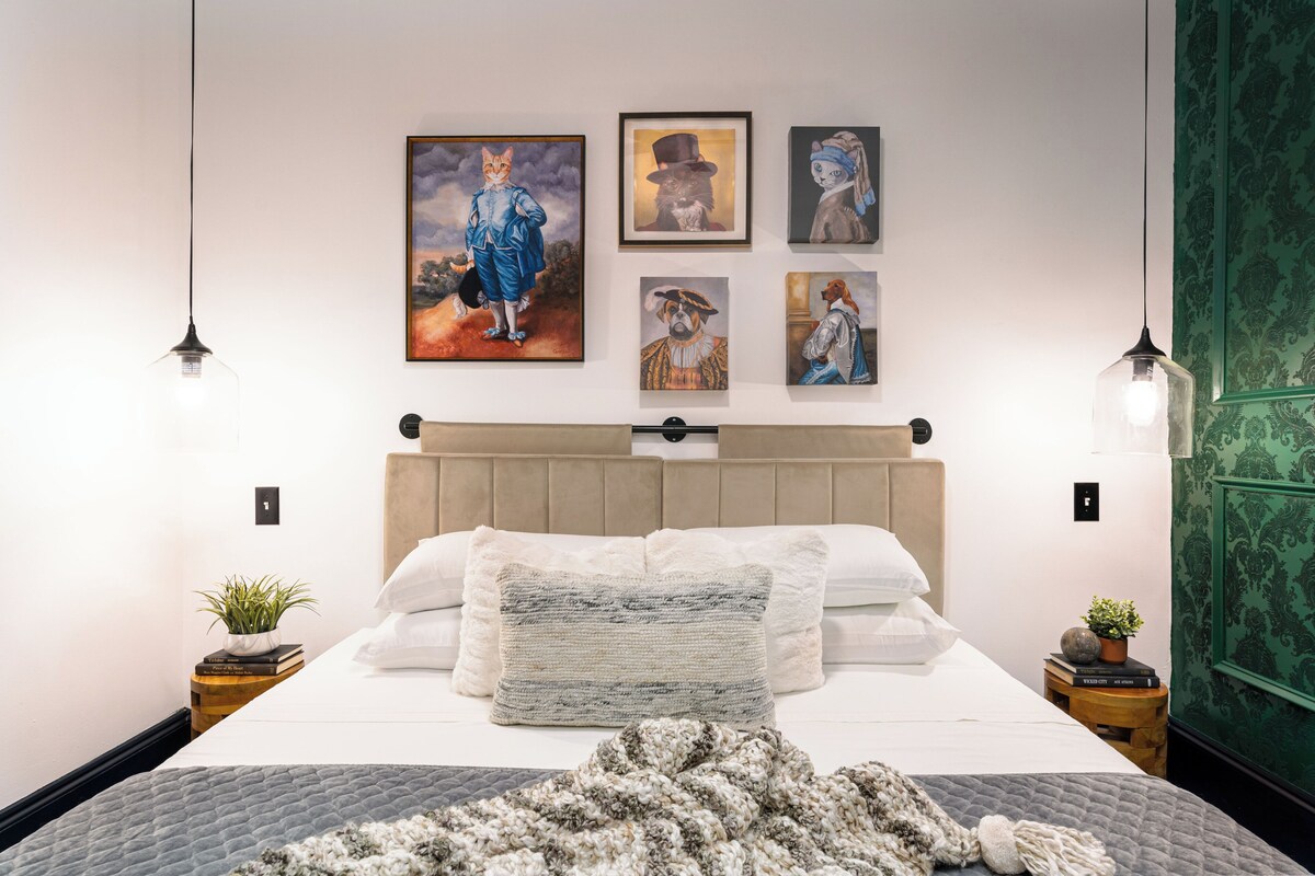 A bedroom with a bed, headboard, pillows, a blanket, wall art, and two bedside tables with plants.