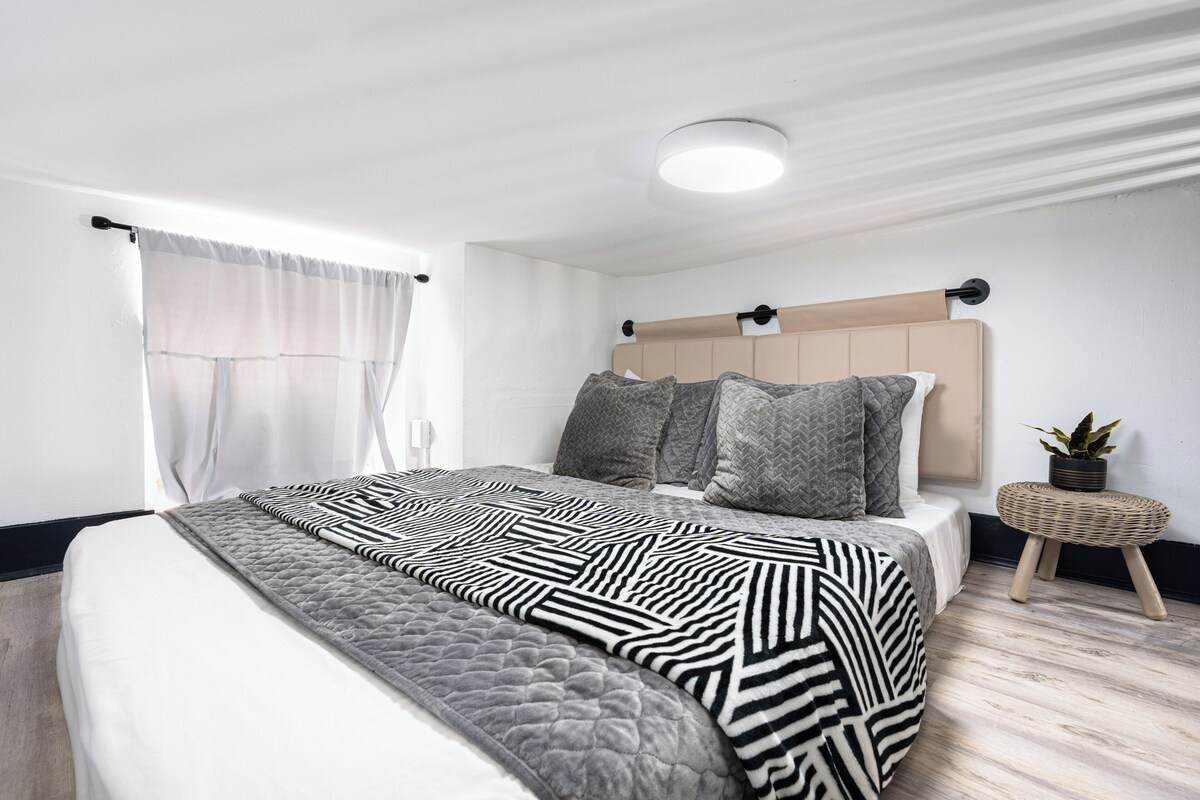 A modern bedroom with a bed, a bedside table with a plant, and a window with curtains.
