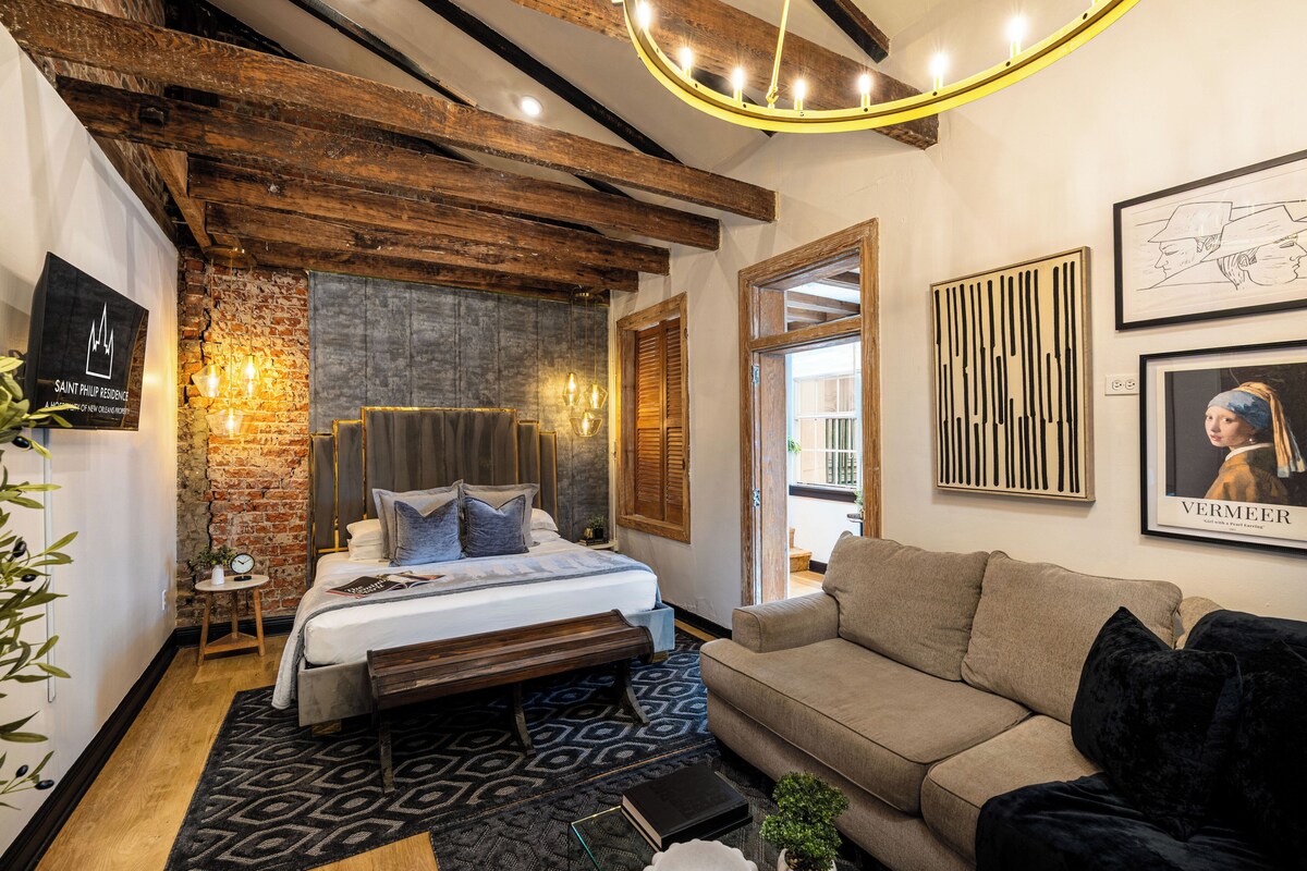 A modern bedroom with exposed wooden beams, a large bed, a sofa, and artwork on the walls.