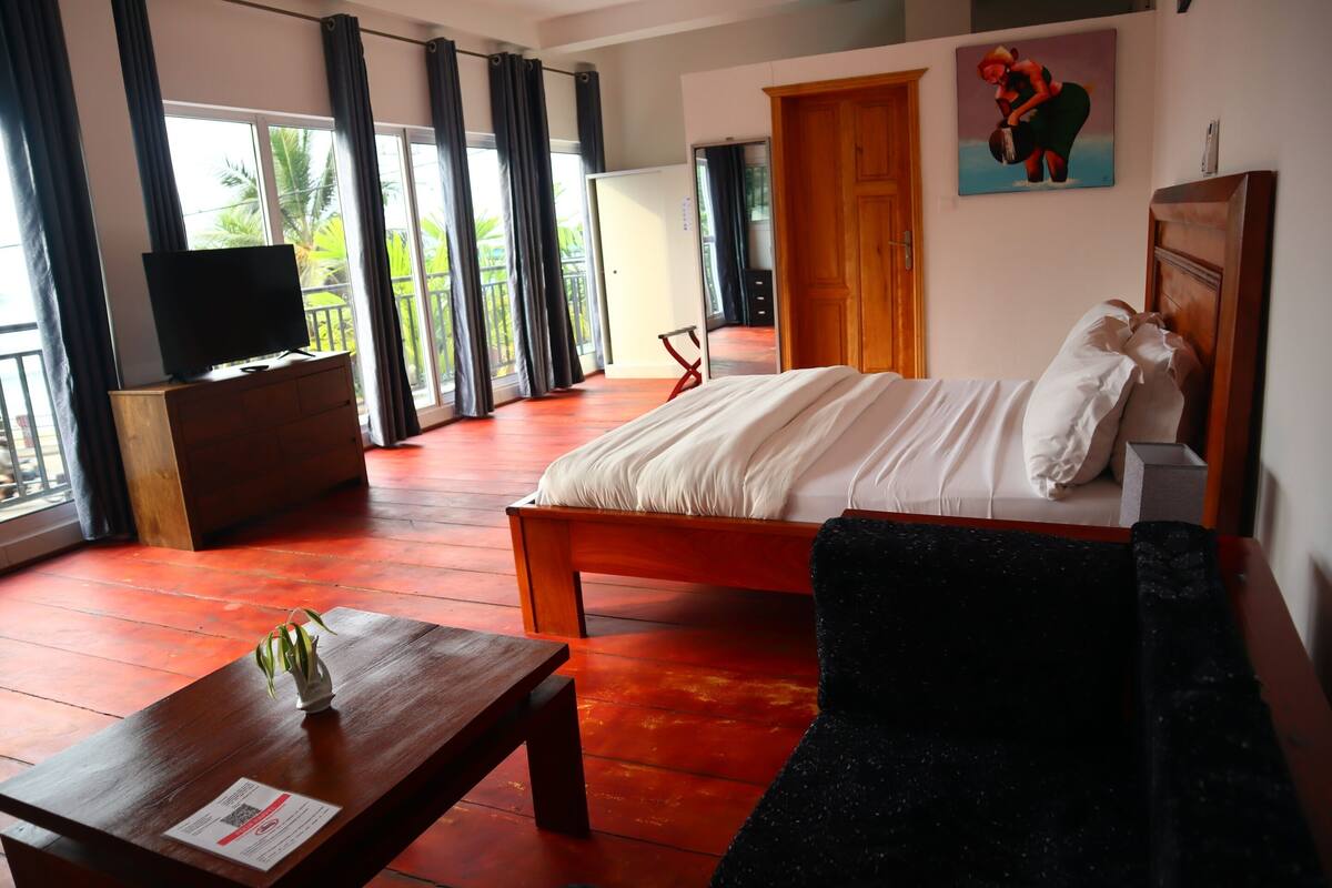 Une chambre d’hôtel avec un lit, un téléviseur posé sur une commode en bois, une petite table avec une plante et un balcon avec des rideaux.
