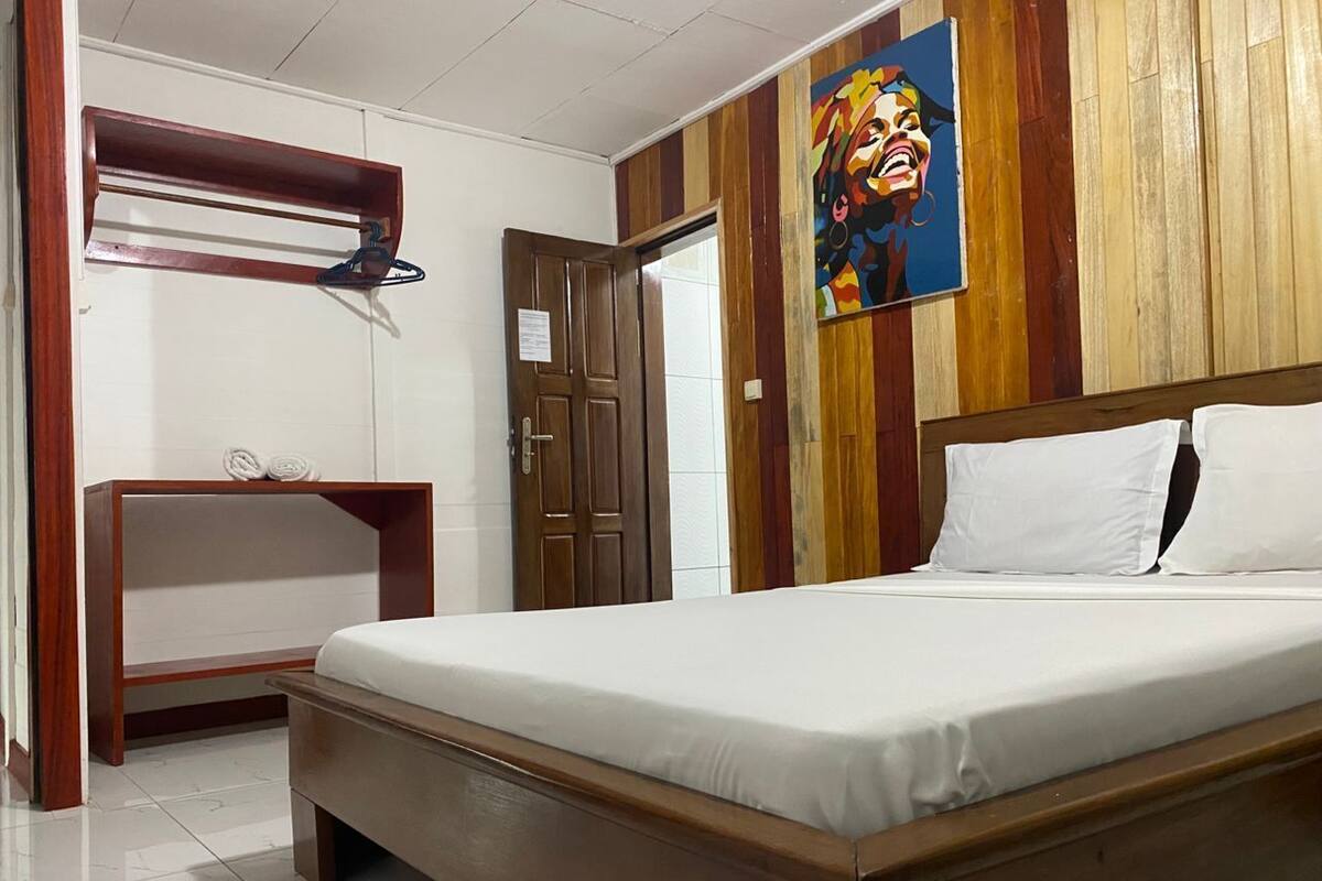 Une chambre d’hôtel avec un lit en bois, une table de chevet en bois, un porte-serviettes et un tableau accroché au mur.