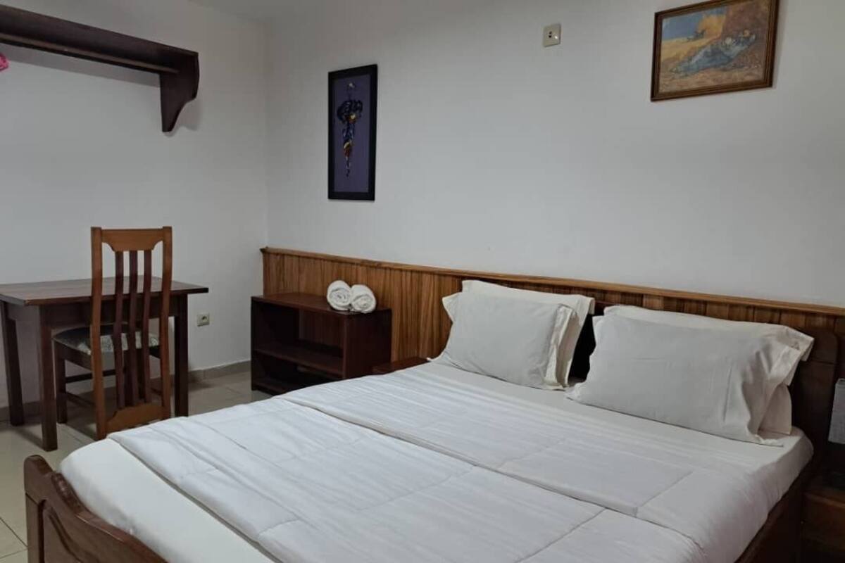 Un lit bien fait, avec des draps blancs, une tête de lit en bois et une table de chevet en bois sur laquelle sont pliées des serviettes.
