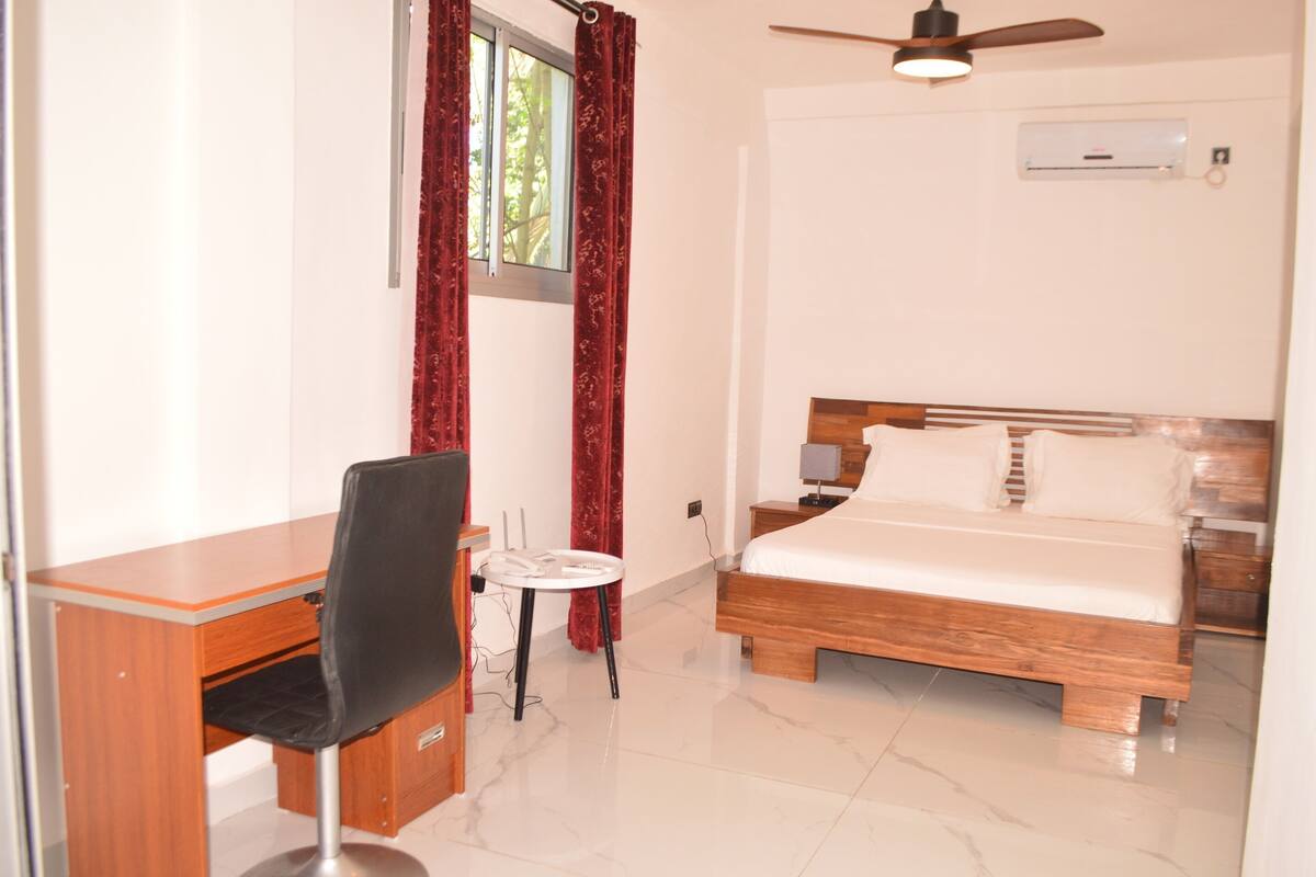 Une chambre à coucher avec un lit en bois, un bureau avec une chaise, une petite table et un ventilateur de plafond.