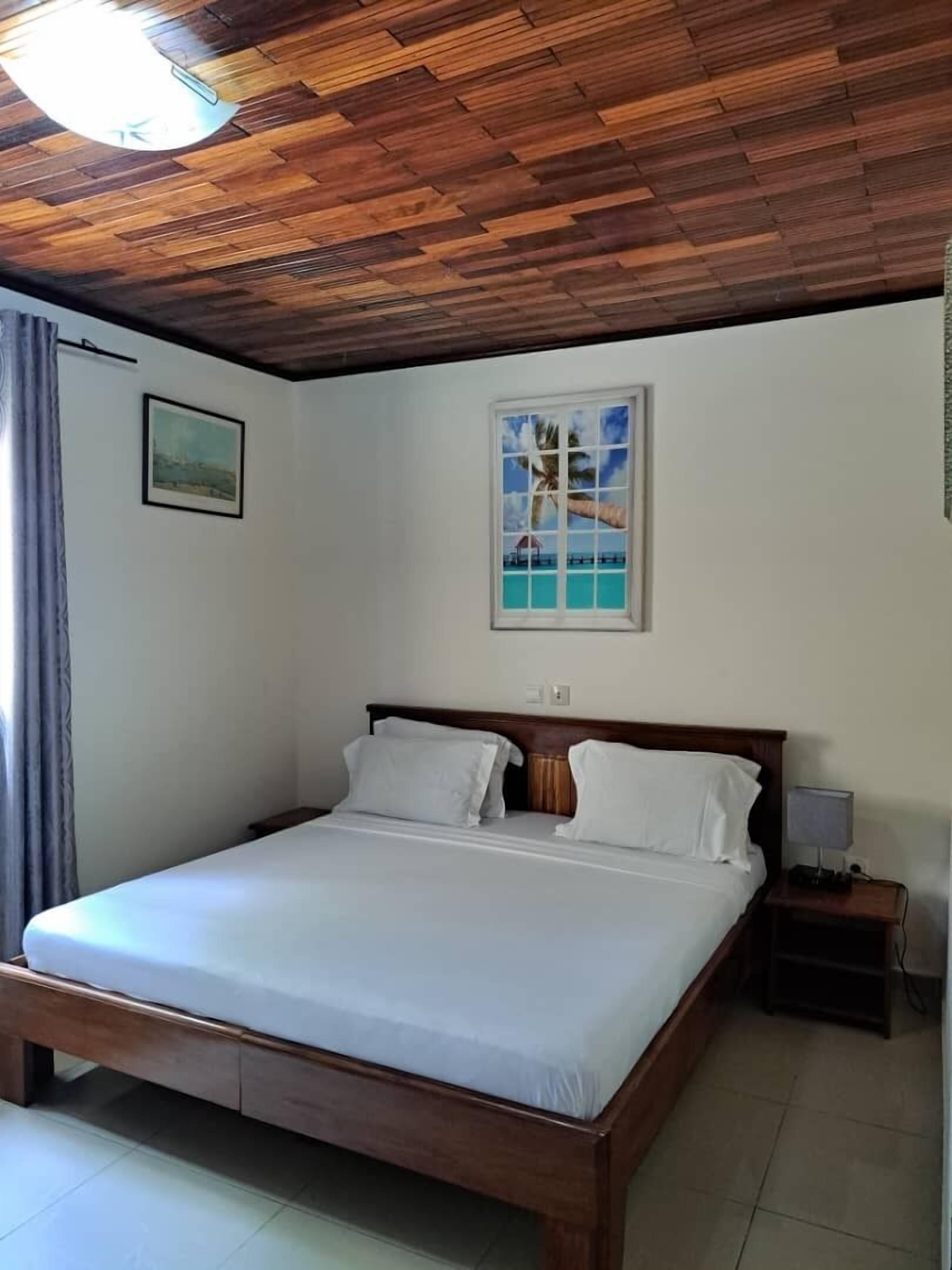 Een enkel bed met houten hoofdeinde, wit beddengoed, een nachtkastje met lamp en een lijst met afbeelding aan de muur.