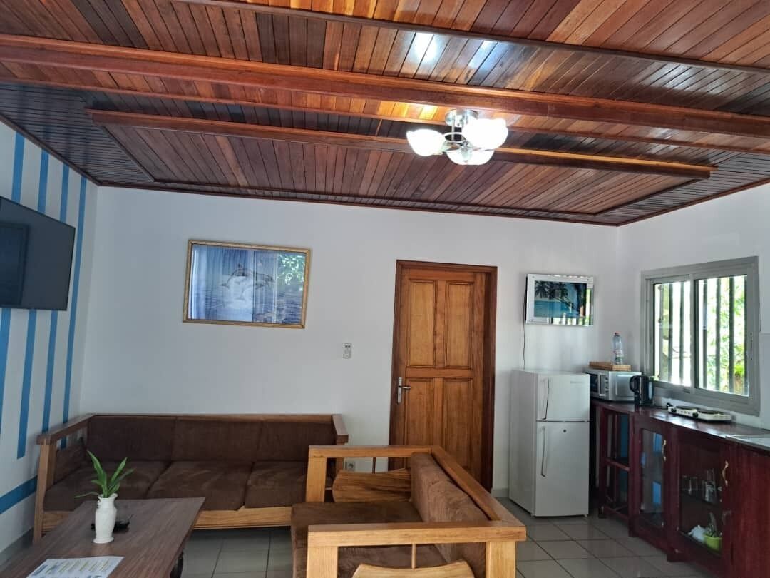 Ein Wohnzimmer mit Holzboden, einem braunen Sofa, einem Holztisch, einer kleinen Pflanze, einem Fernseher und einem Kühlschrank.