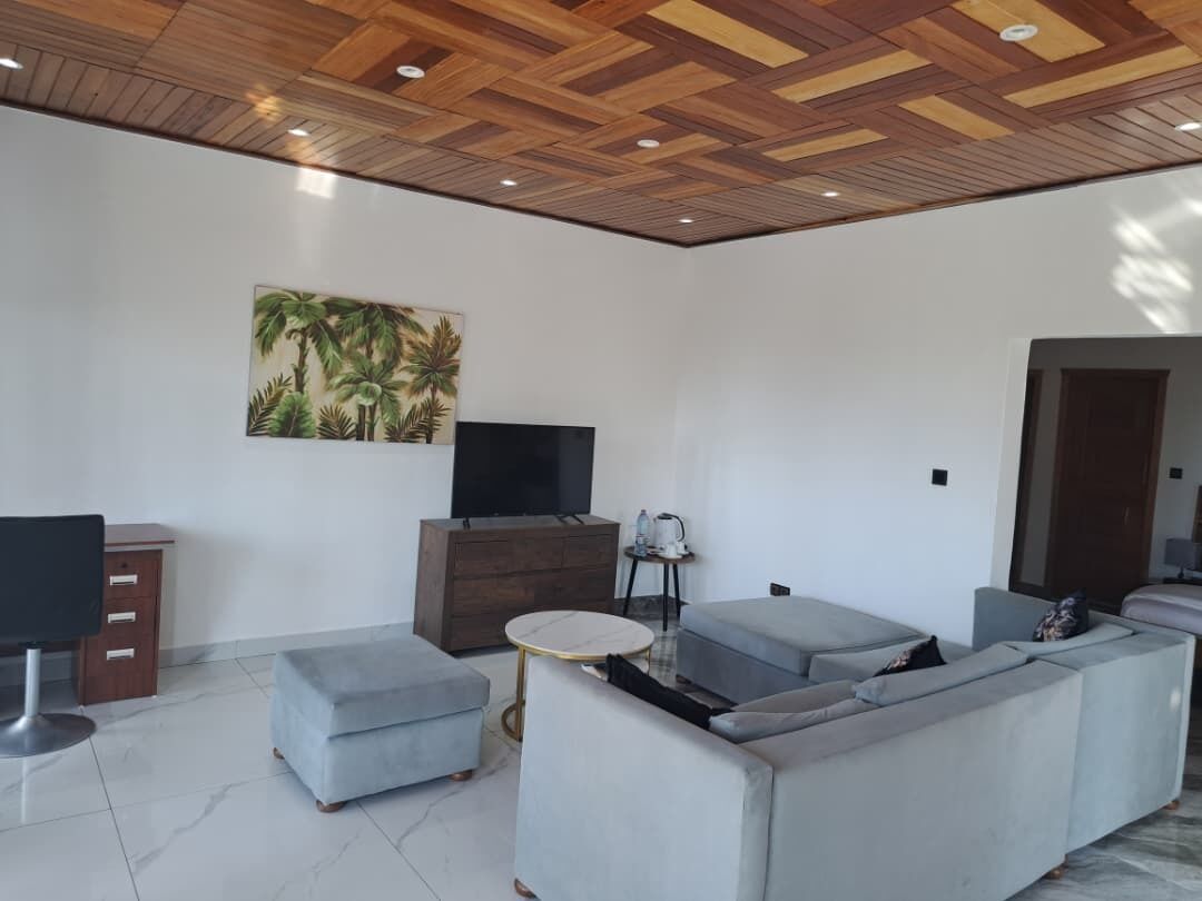 A modern living room with a wooden ceiling, a large sofa, a TV, a small table, and a painting of palm trees.