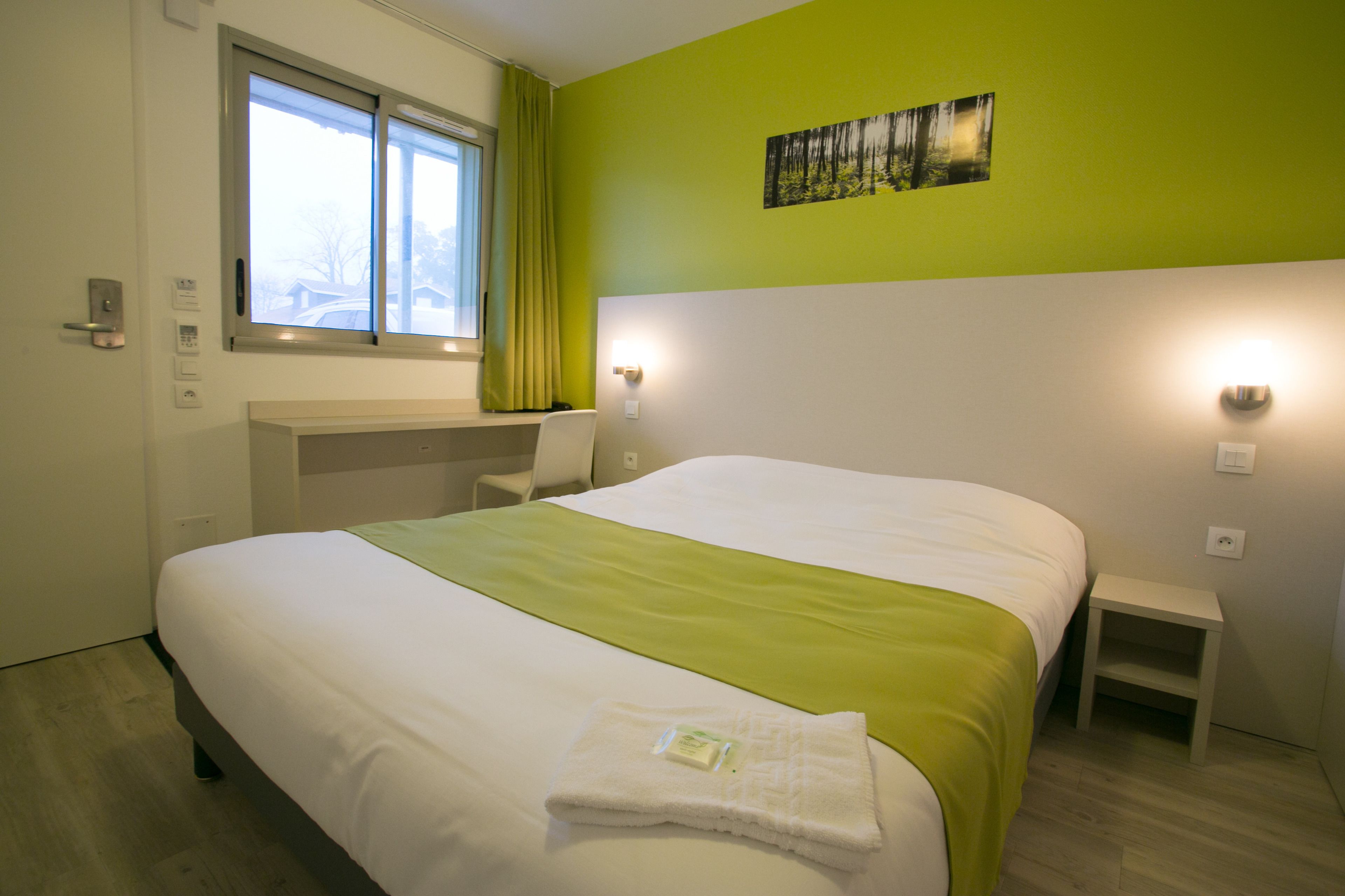 A hotel room with a bed, a window, a nightstand, and a wall-mounted light fixture.