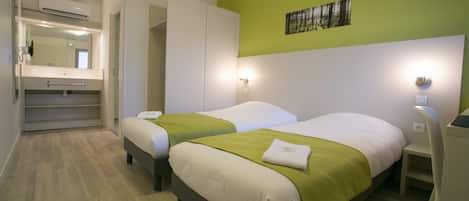 A hotel room with two beds, a green accent wall, a painting, and a bathroom with a mirror and towel rack.
