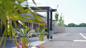 Un espace de stationnement avec un bâtiment en arrière-plan, un bac à fleurs vert contenant des plantes et un ciel dégagé.