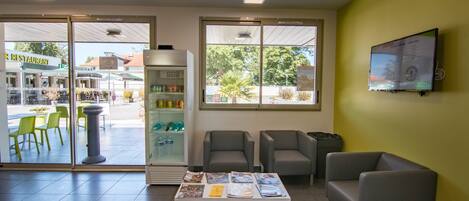 A reception area with a reception desk, two armchairs, a small table with brochures, a refrigerator, and a TV on the wall.