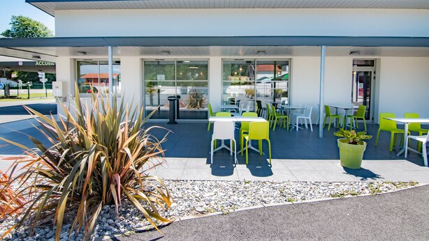 Un espace extérieur aménagé en terrasse, avec des tables et des chaises, un pot de fleurs et l’entrée d’un bâtiment.