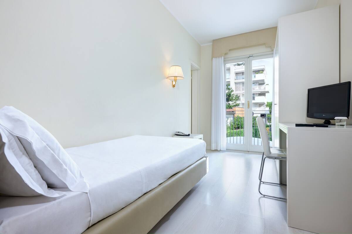Une chambre d’hôtel moderne, équipée d’un lit, d’un bureau avec une télévision, d’une lampe et donnant sur le balcon extérieur.
