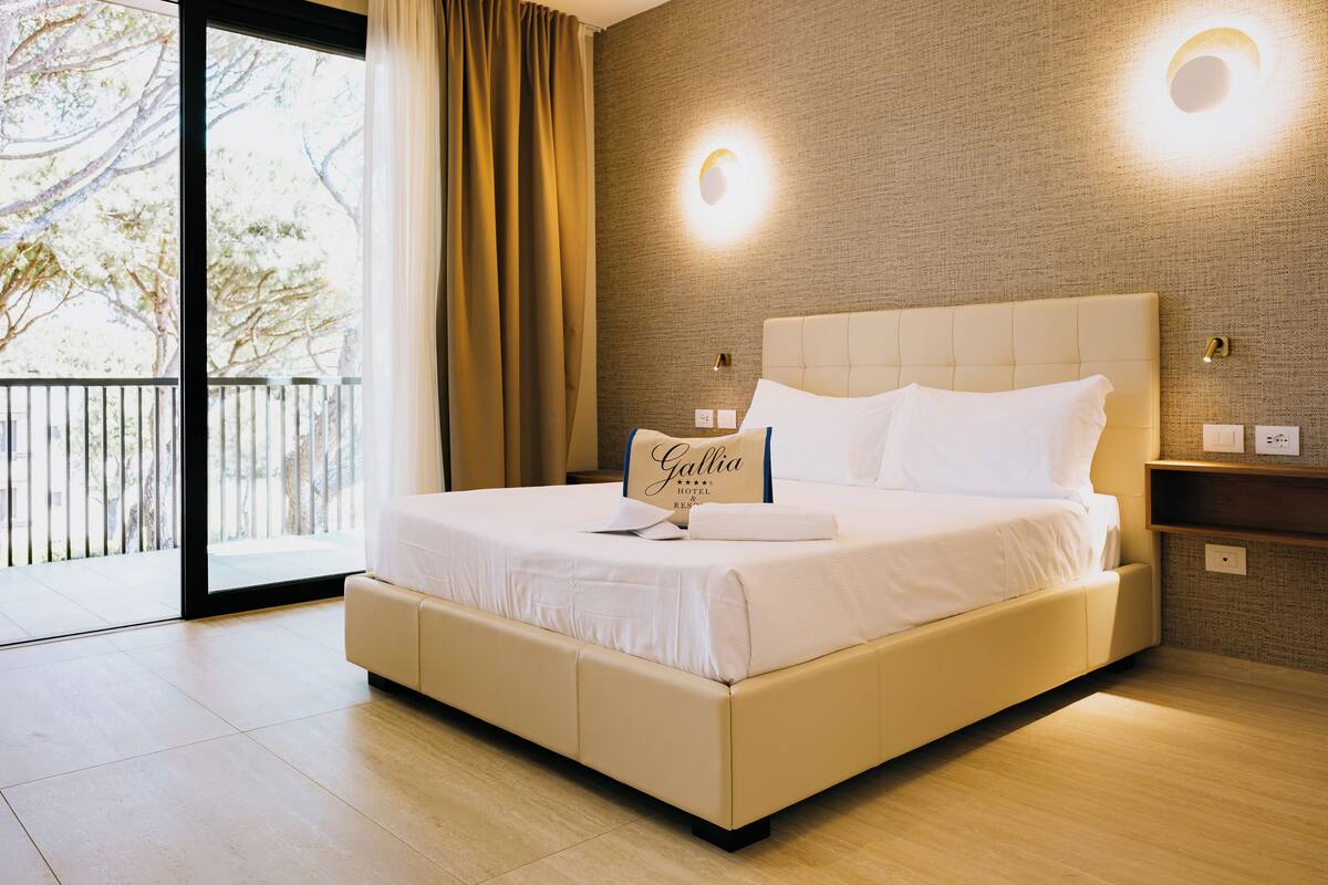 Une chambre d’hôtel moderne avec un grand lit, une table de chevet, une lampe fixée au mur et une vue sur l’extérieur.