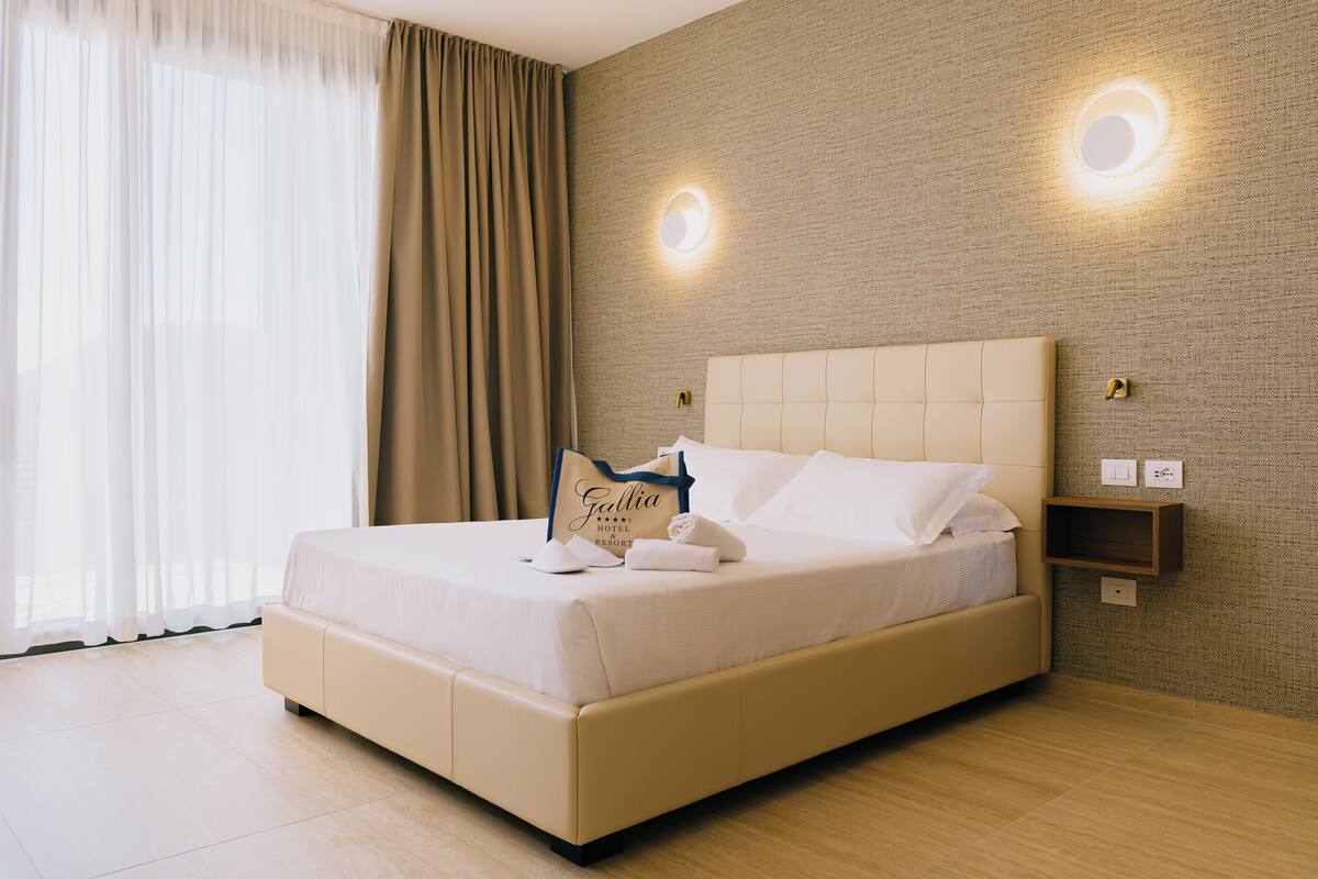 Une chambre d’hôtel avec un grand lit, une table de chevet, une étagère fixée au mur et une fenêtre avec des rideaux.