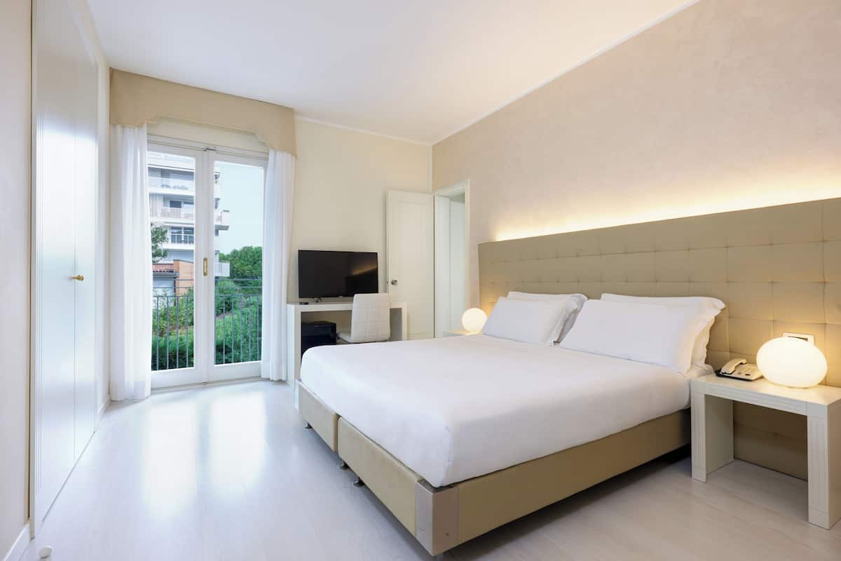 Une chambre d’hôtel moderne dotée d’un grand lit, de tables de chevet, d’une télévision et d’un balcon offrant une vue sur les bâtiments et la verdure.
