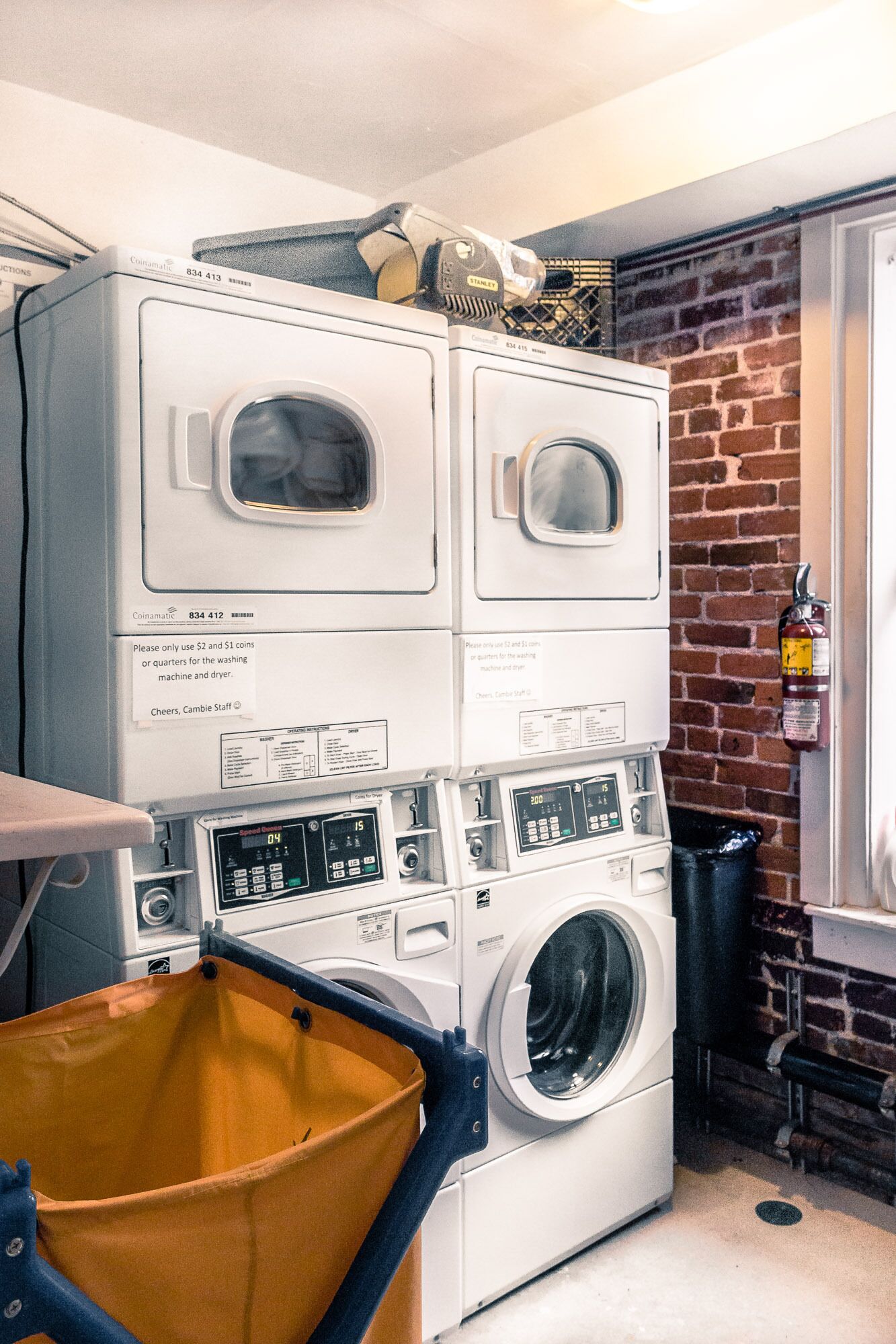 A laundry room with stacked washing machines and dryers, a fire extinguisher, and a yellow laundry basket.