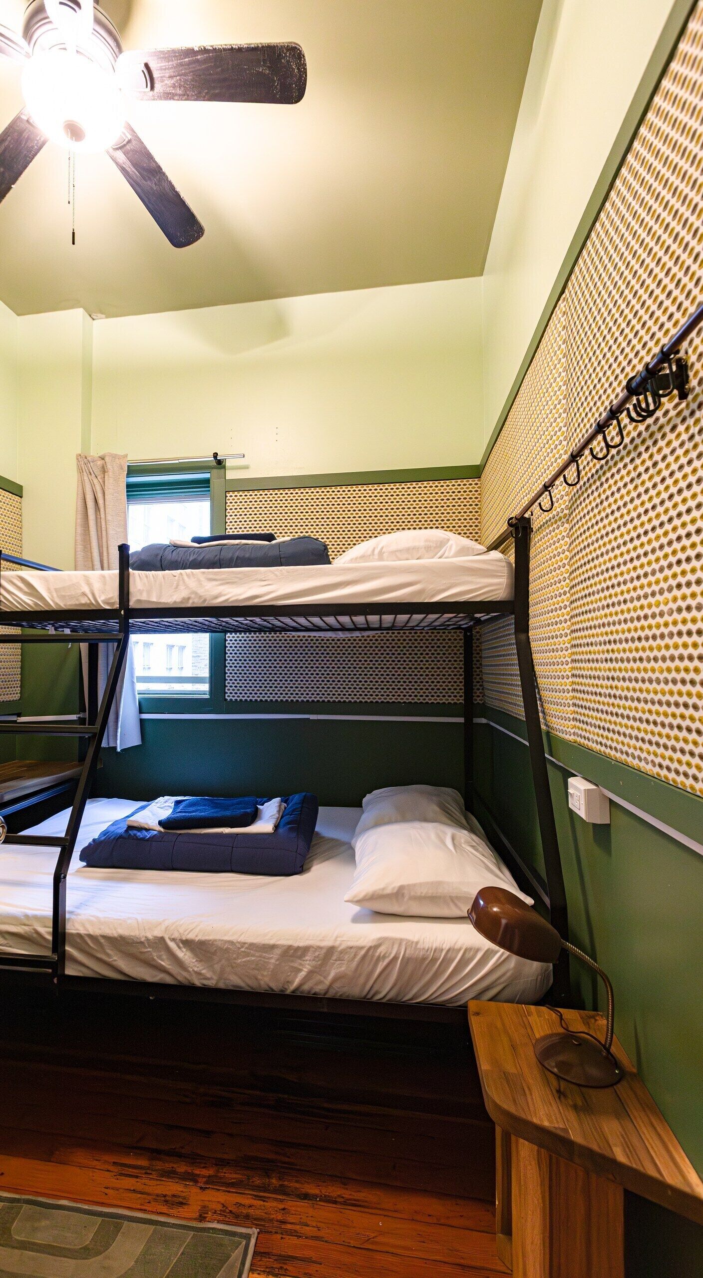 Een slaapkamer met stapelbed, plafondventilator, raam met gordijnen en een groene wand met een geprint paneelontwerp.