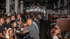 A crowded bar with patrons seated at tables, a television screen displaying a boxing match, and a brick wall interior. - Cambie Downtown Hostel (Vancouver)