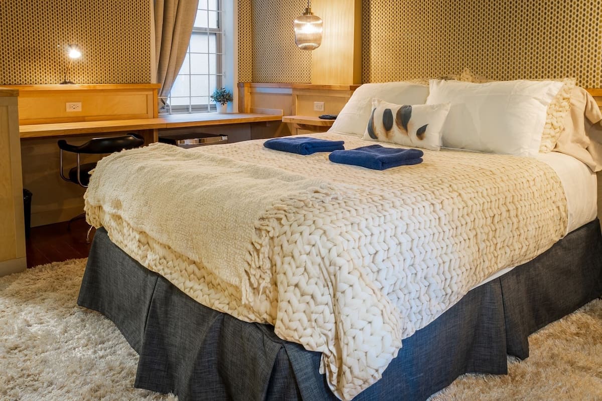 Un lit bien fait, avec une couverture à motifs et des oreillers, un bureau avec une lampe et une fenêtre avec des rideaux.