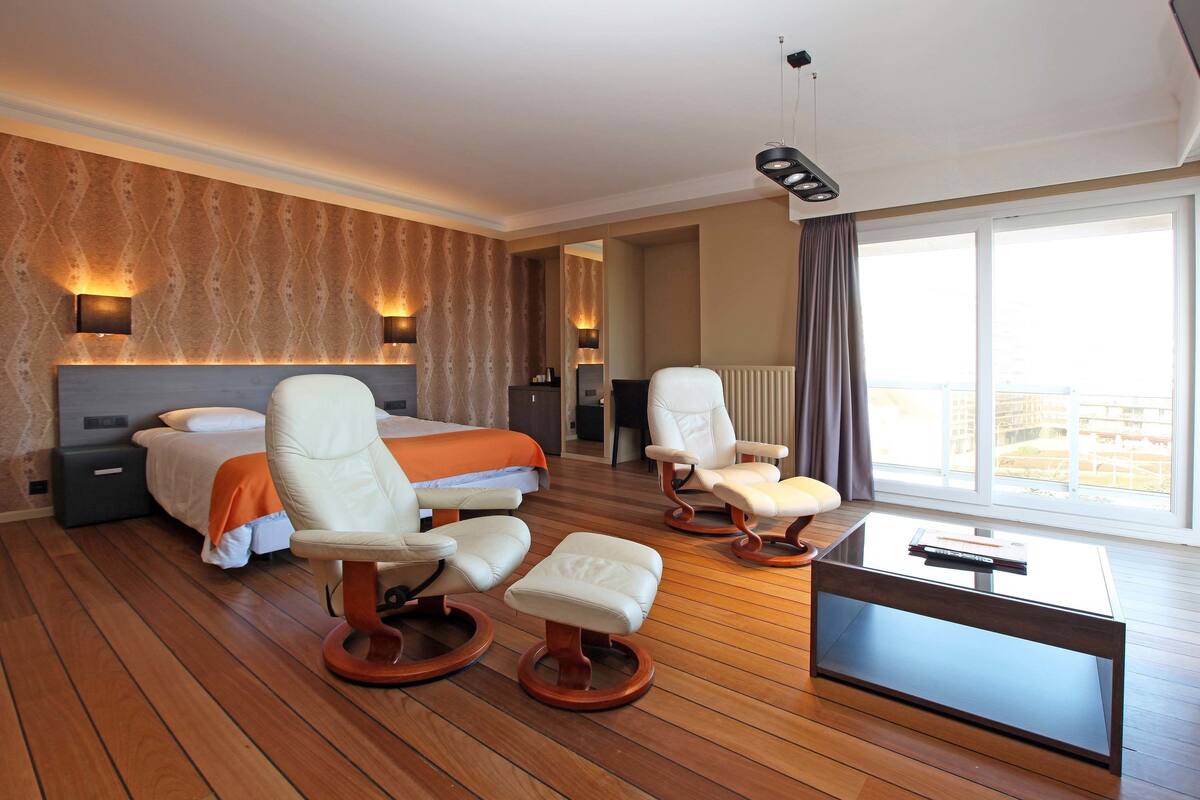 A modern hotel room with a large bed, two white armchairs, a glass coffee table, and a view of the city through large windows.