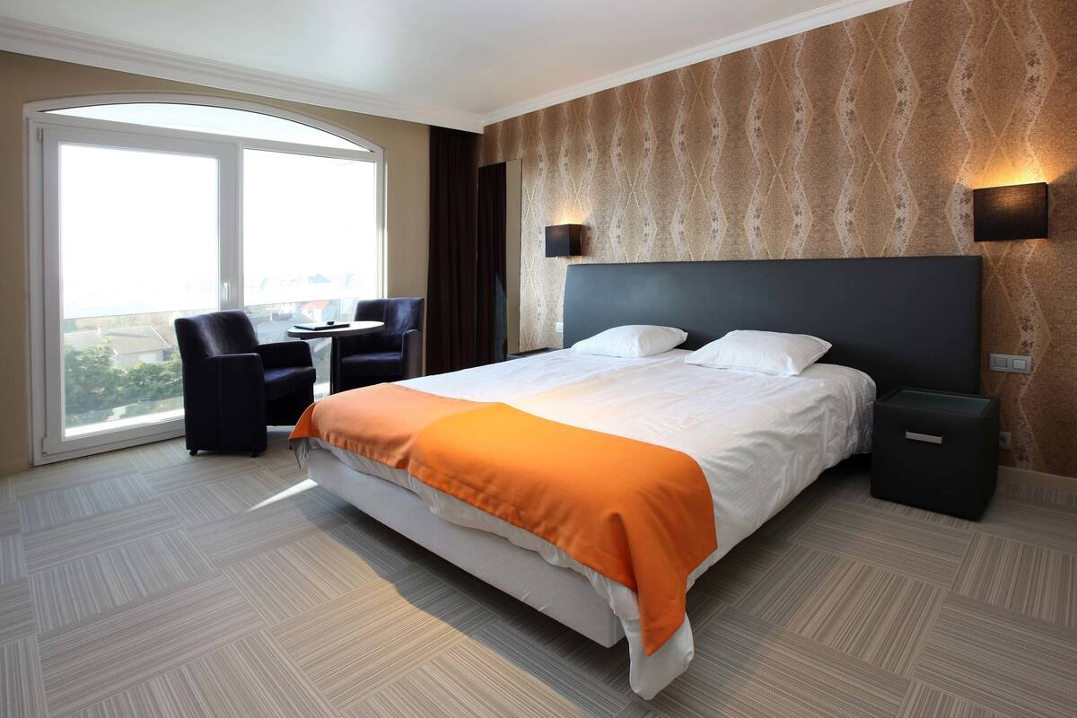 A hotel room with a large bed, a desk, and a chair. There is a window providing a view of the outside.