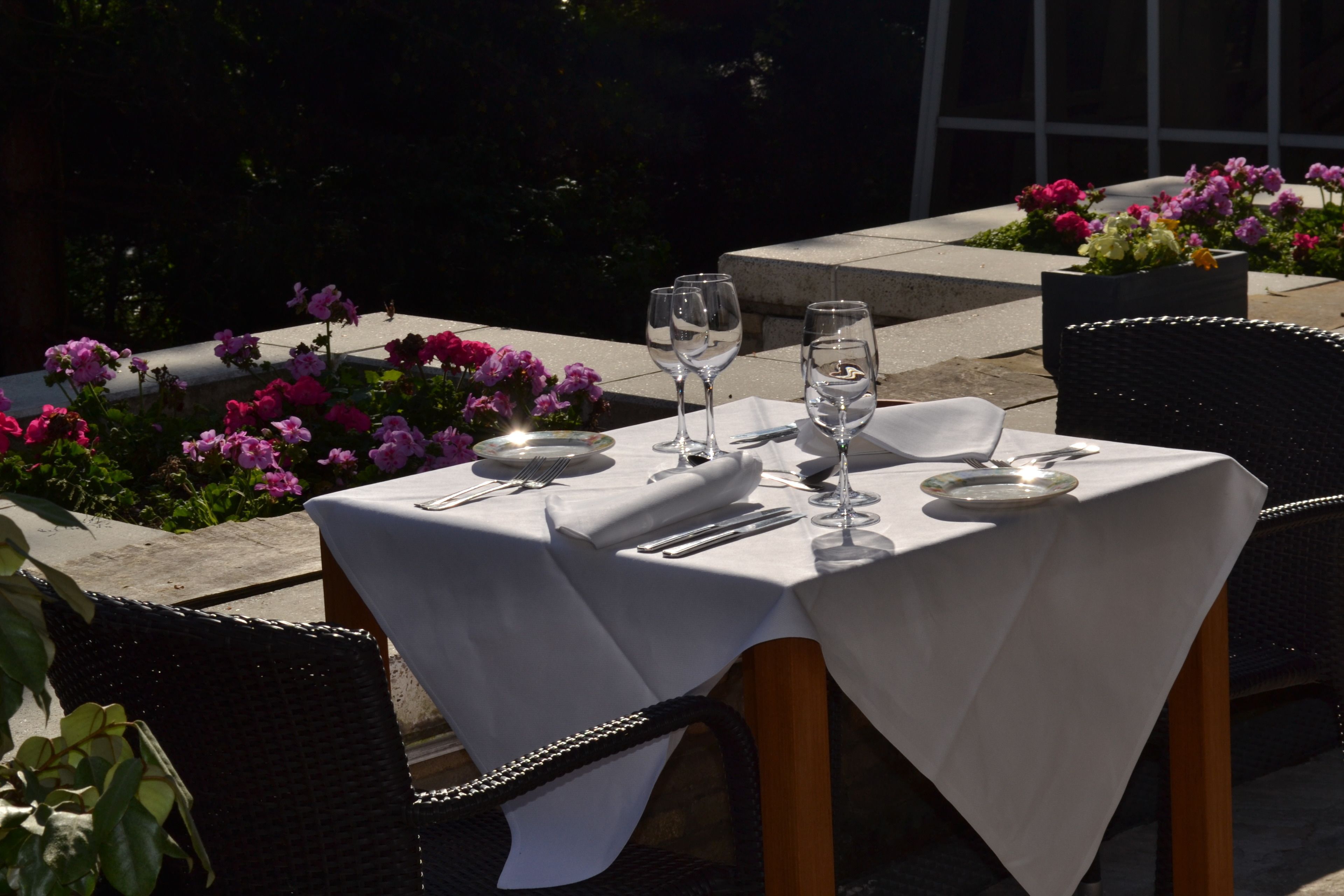 Mesa de comedor exterior con mantel blanco, platos, cubiertos y vasos, rodeada de macetas con plantas y flores.