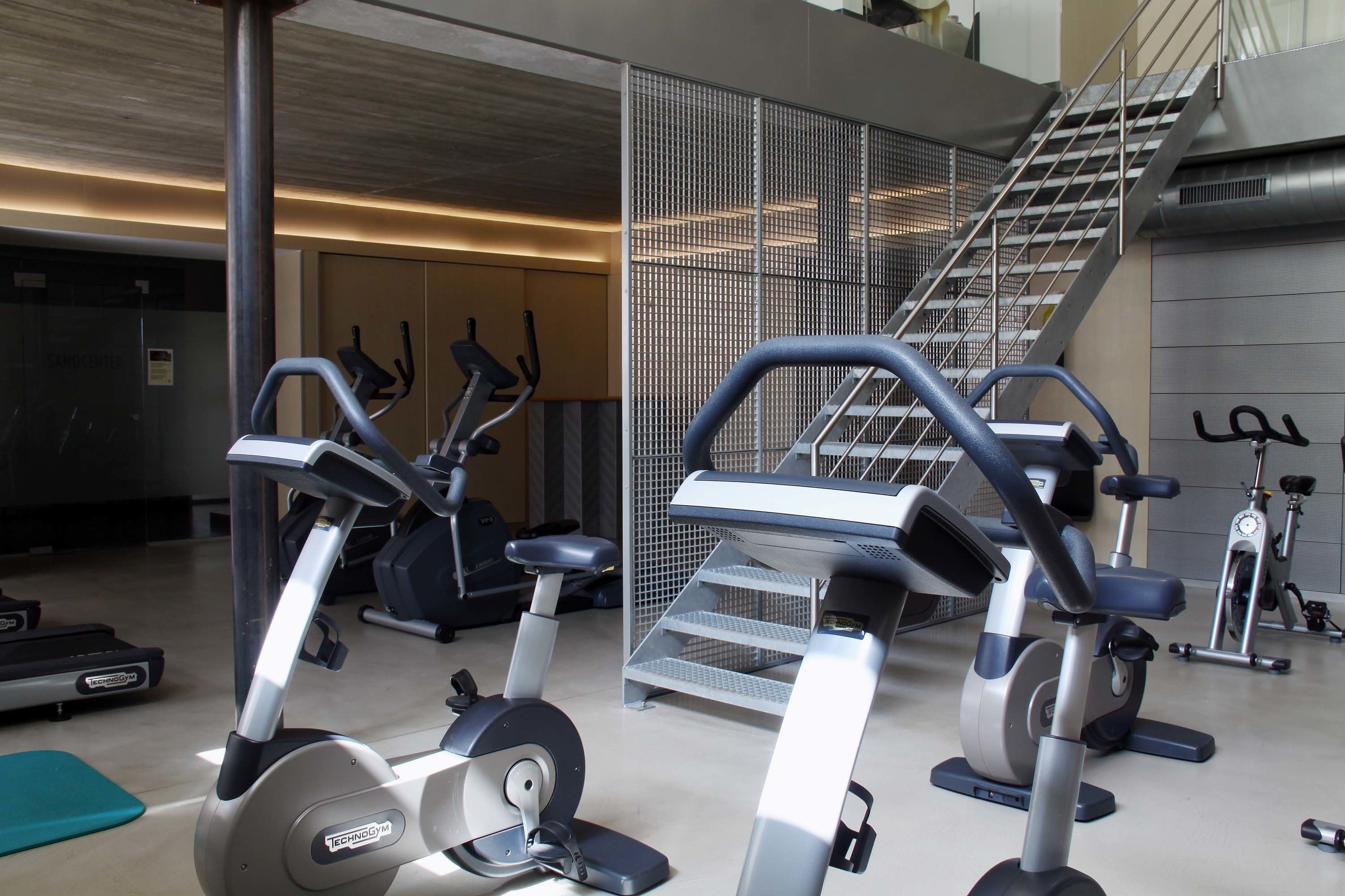 Een sportschool met diverse fitnessapparaten, waaronder loopbanden en elliptische trainers, gerangschikt in een ruime ruimte met een trap naar een verdieping.