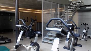 Een sportschool met diverse fitnessapparaten, waaronder loopbanden en elliptische trainers, gerangschikt in een ruime ruimte met een trap naar een verdieping.