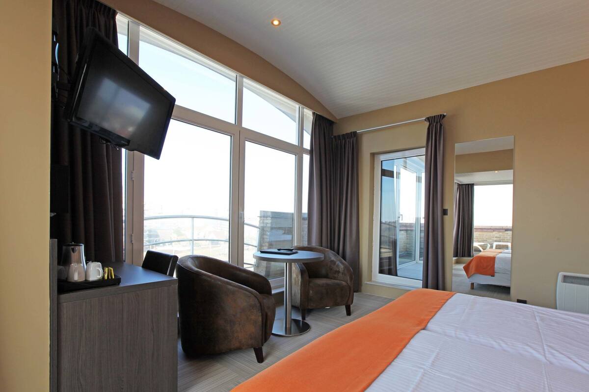 A hotel room with a bed, two armchairs, a small table, a TV, and a balcony with a view.