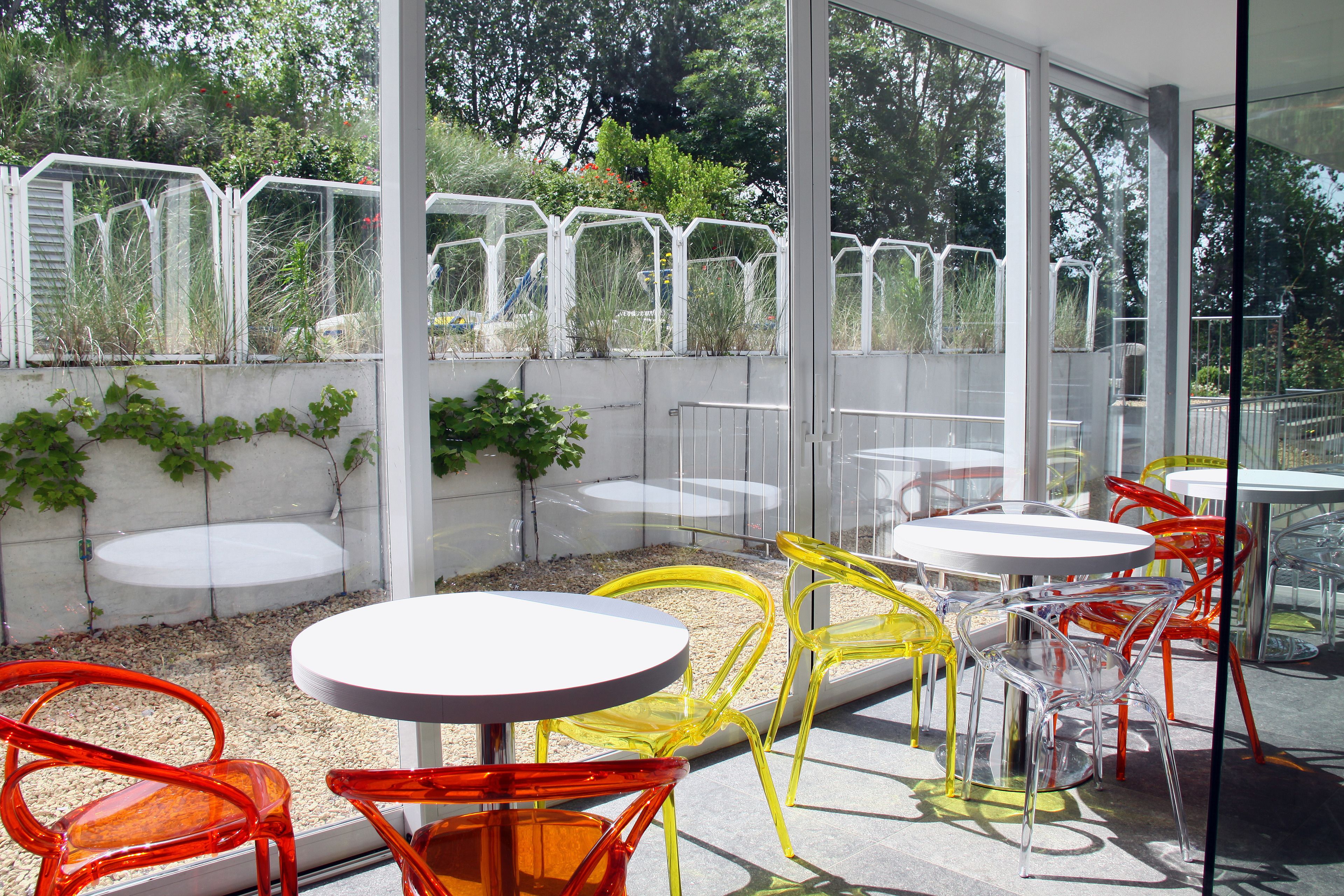 Un patio exterior con mesas y sillas redondas, rodeado por un recinto de vidrio con vistas a follaje.