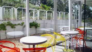 Un patio exterior con mesas y sillas redondas, rodeado por un recinto de vidrio con vistas a follaje.