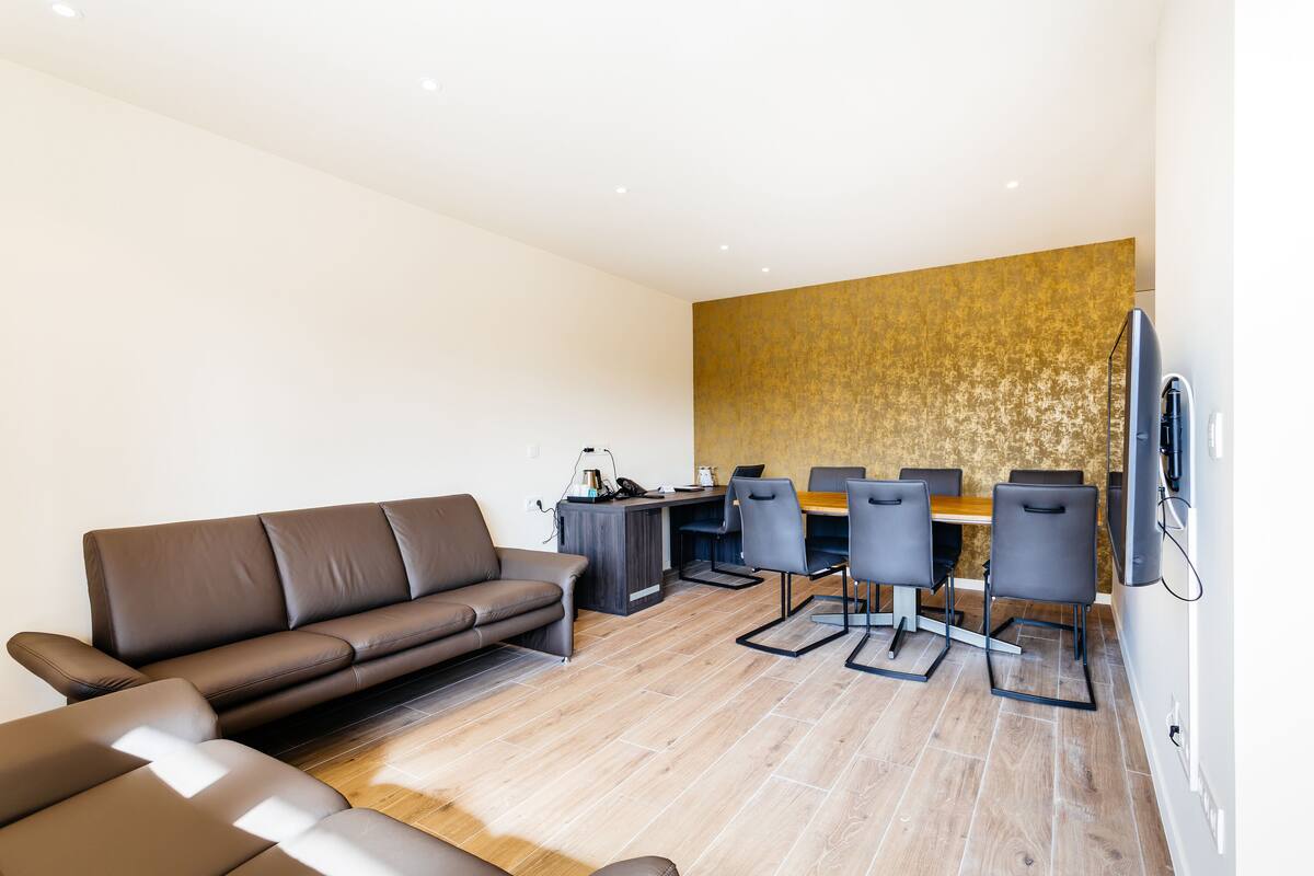 A modern living room with a brown sofa, a dining area with a wooden table and chairs, and a large flat-screen TV mounted on the wall.