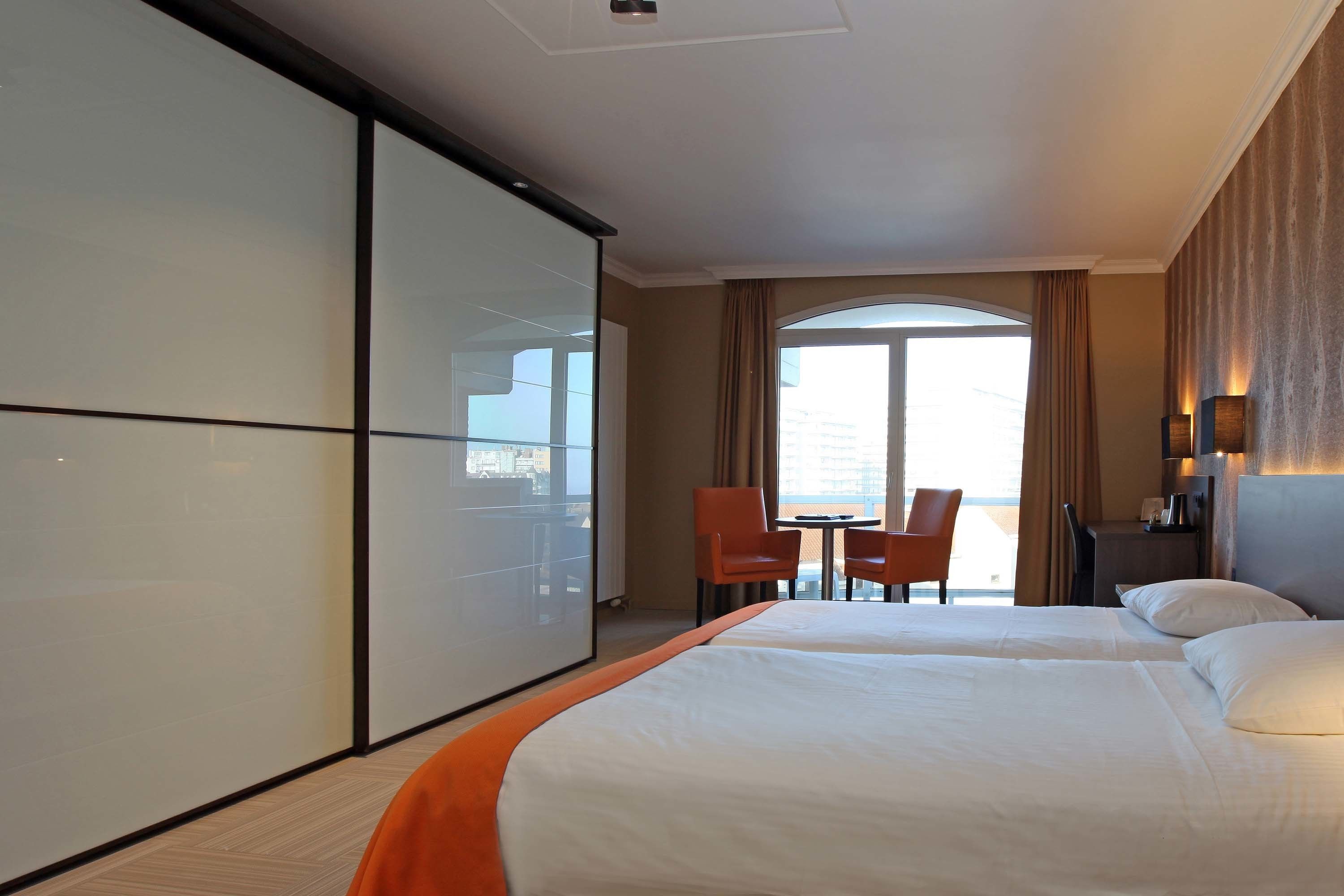 A hotel room with two beds, a sliding door, a desk with a chair, and a view of the city through a large window.