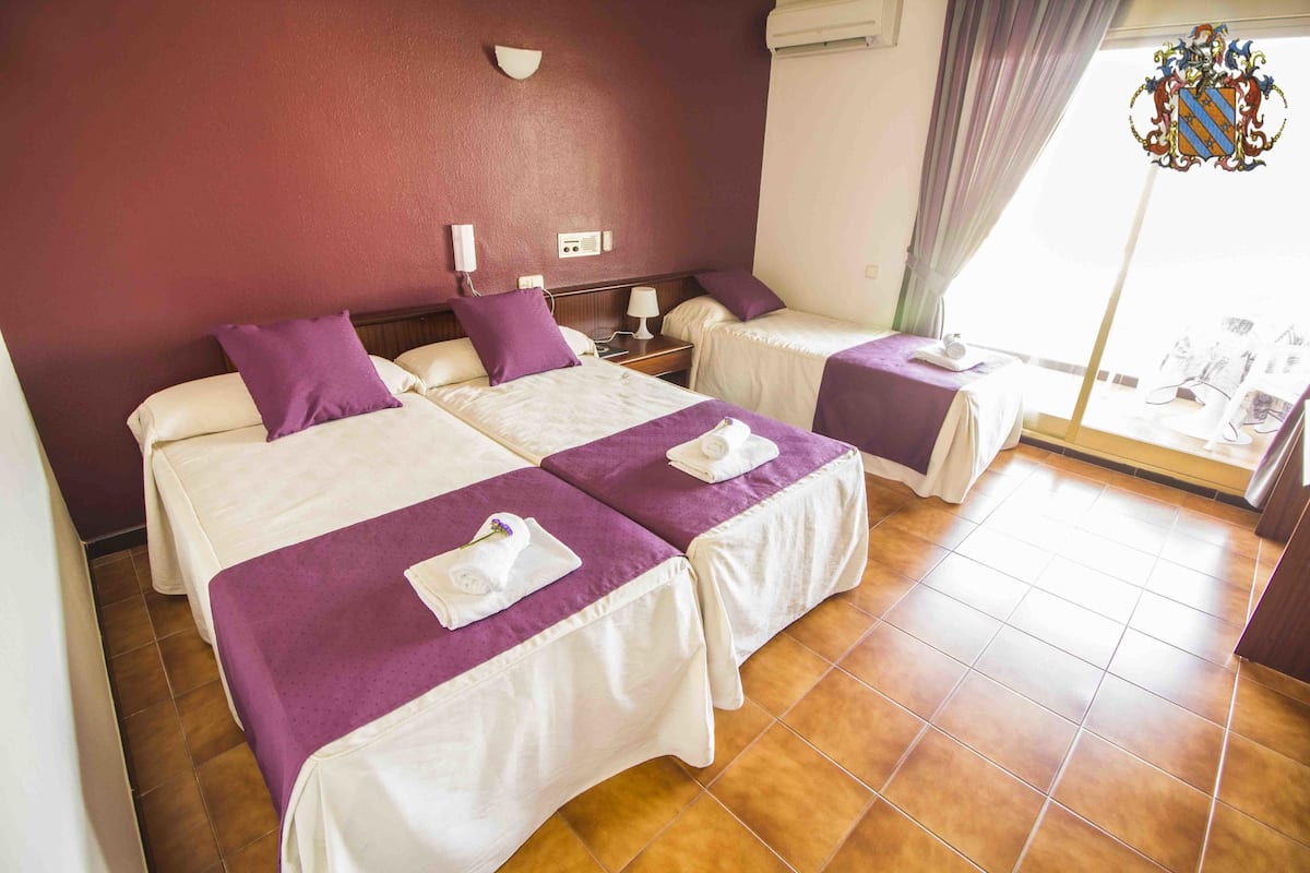 A hotel room with two beds, each with a purple bedspread and white linens, a nightstand with a lamp, a wall-mounted phone, and a window with curtains.