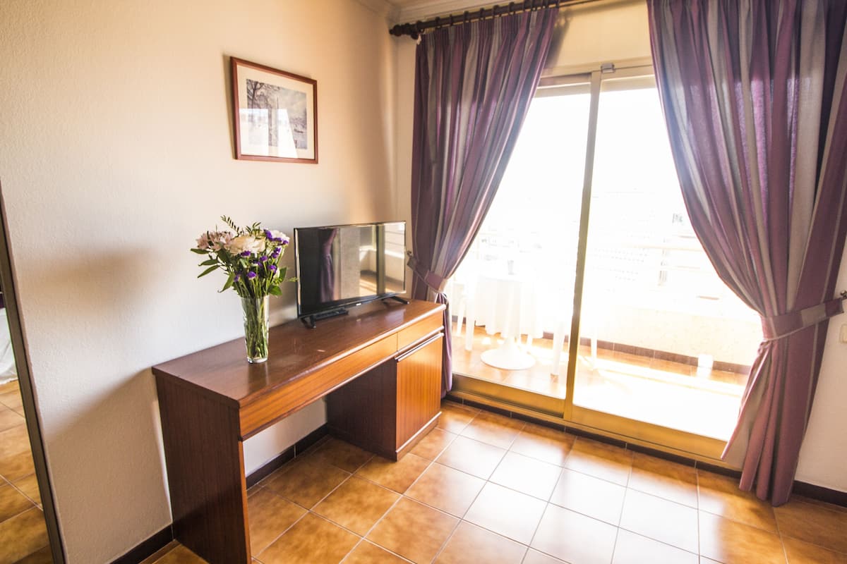 A room with a wooden desk, a flat-screen TV, a vase with flowers, and a sliding glass door with curtains.