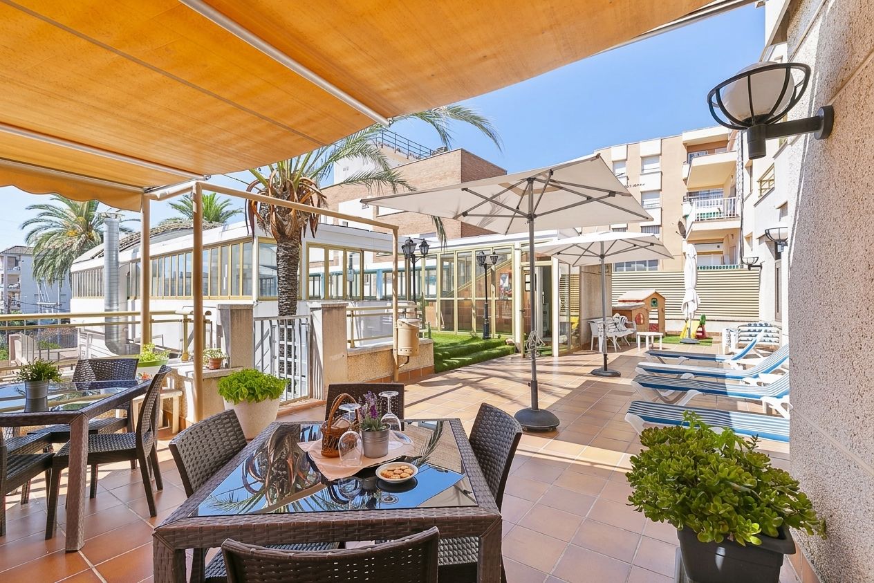 An outdoor patio area with tables and chairs, a glass-top table, potted plants, and umbrellas providing shade.