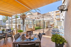 An outdoor patio area with tables and chairs, a glass-top table, potted plants, and umbrellas providing shade. - HOTEL VICTORIA (Calafell)